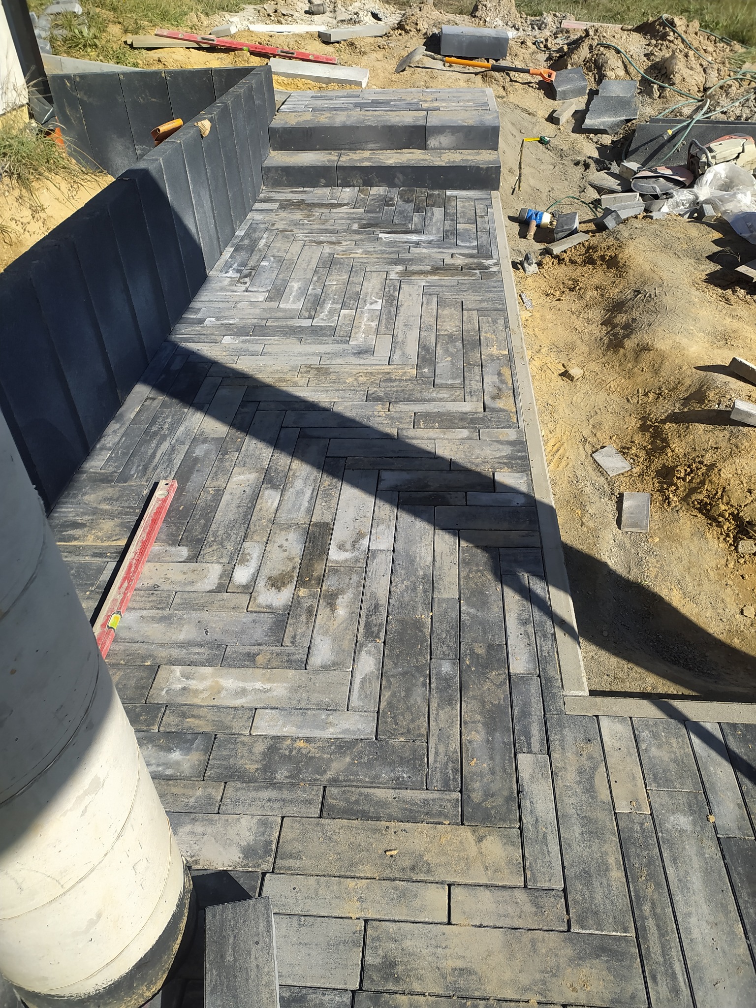 Układanie szarej kostki brukowej w jodełkę, widoczny poziomica, krawężnik i przygotowane podłoże.