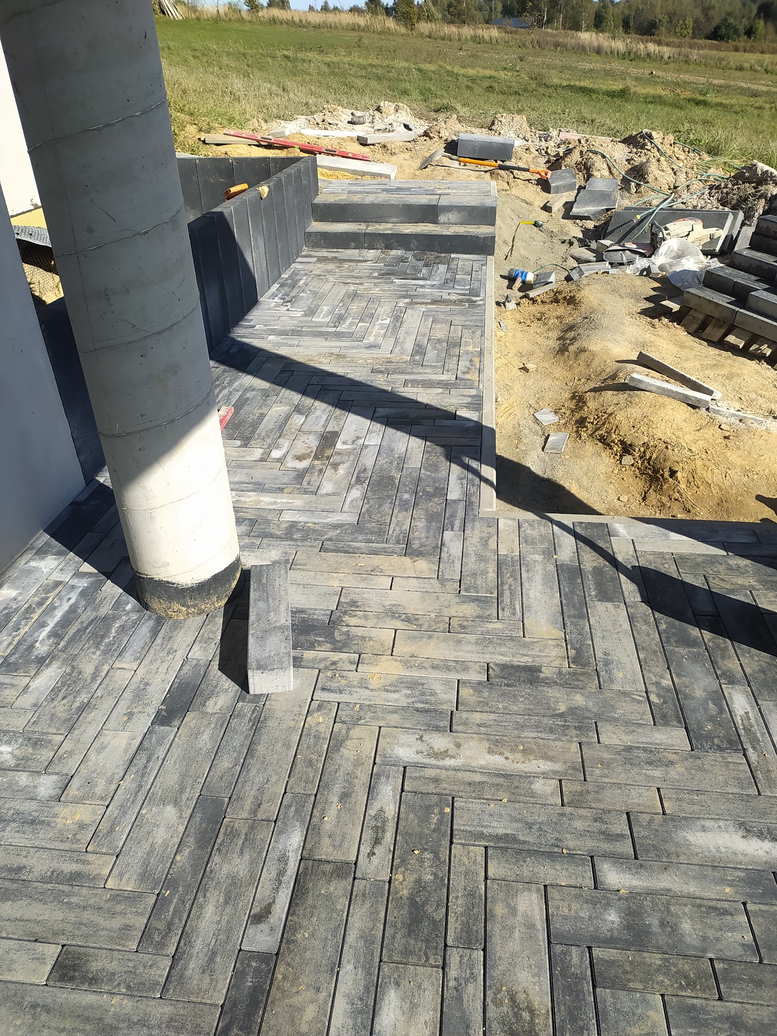 Ułożona kostka brukowa w jodełkę przy betonowym słupie, widoczne schody z kostki i przygotowany teren z narzędziami w tle.