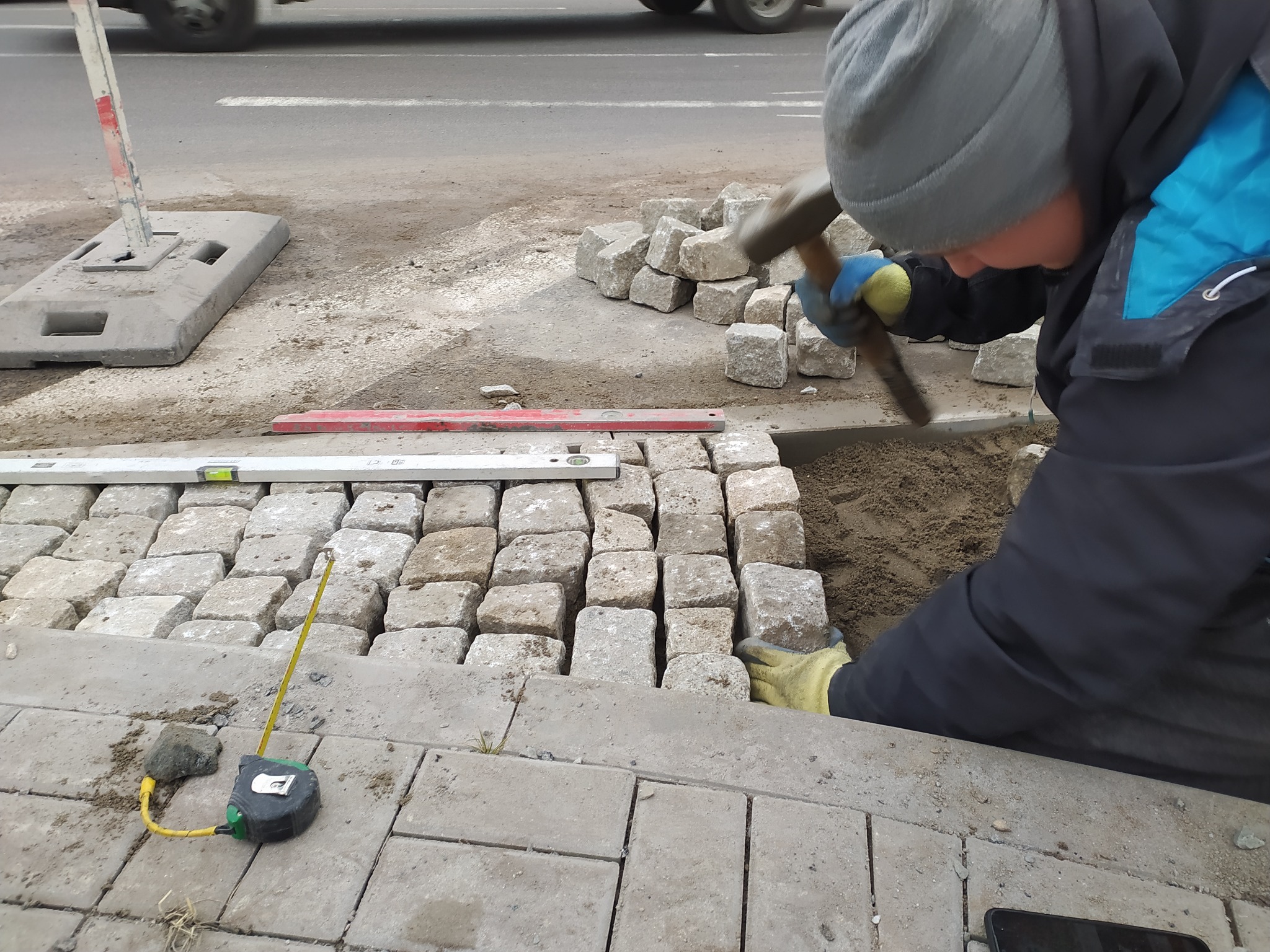 Pracownik w rękawicach układa kostkę granitową przy drodze, używając młotka do osadzania, obok leżą poziomica i zwinięta miarka.
