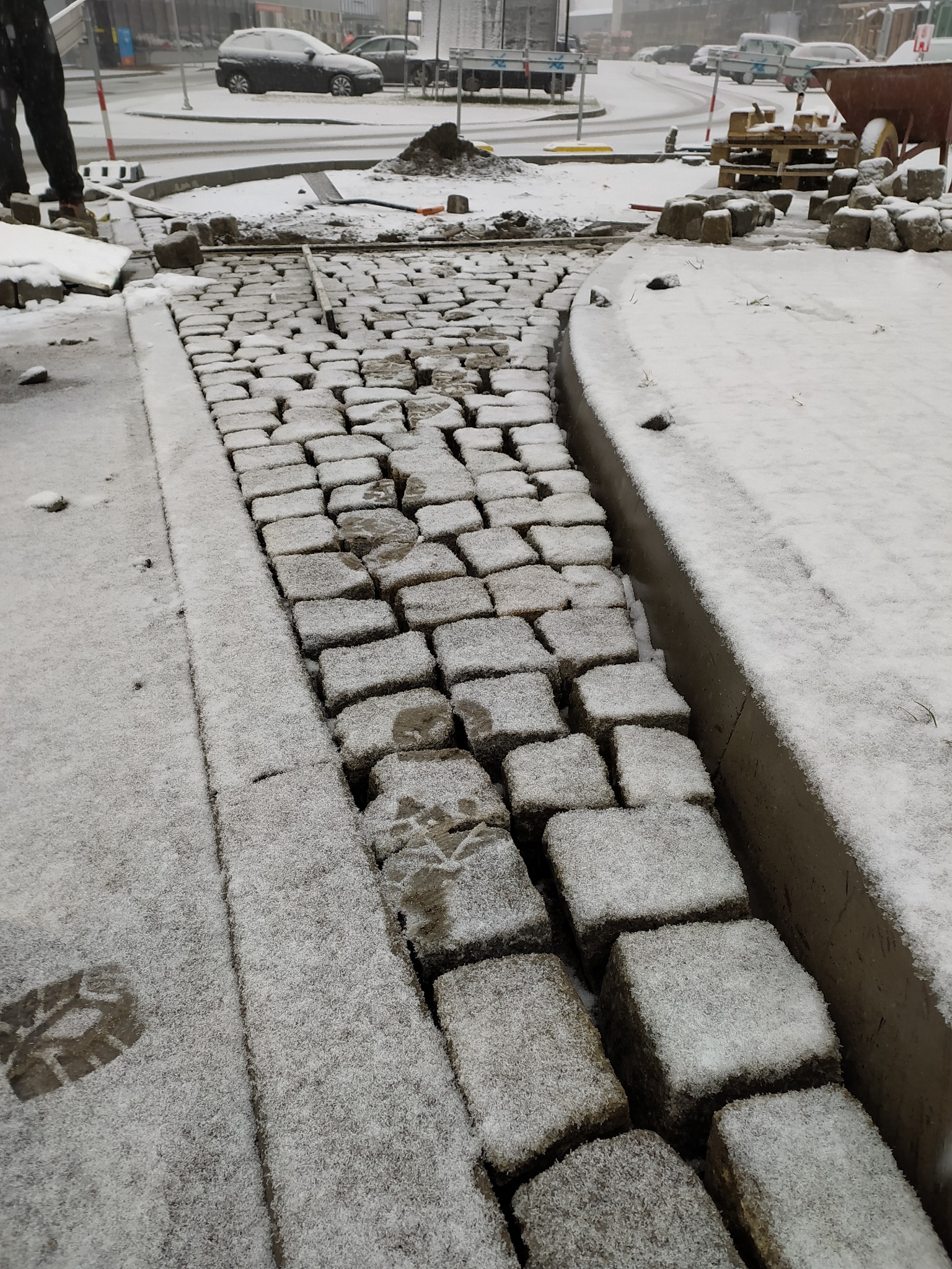 Świeżo ułożona ścieżka z kostki granitowej pokryta cienką warstwą śniegu, widoczne odciski butów na kostce i obok niej, palety z kostką w tle.