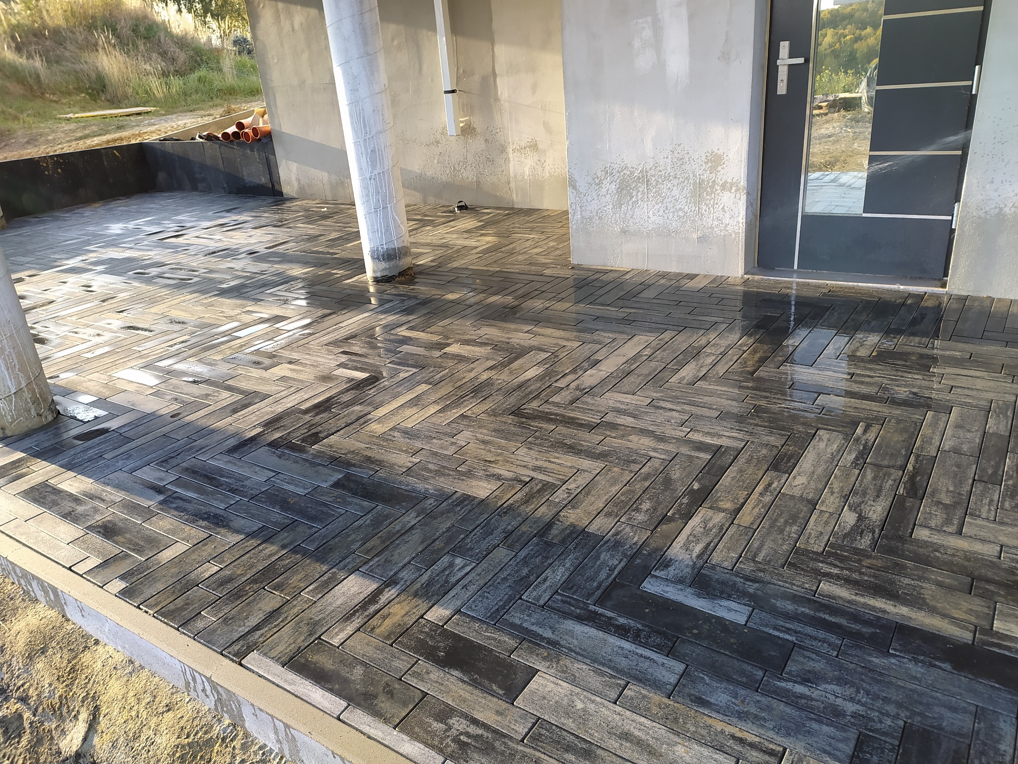 Ułożona, ciemnoszara kostka brukowa w jodełkę na tarasie domu w trakcie budowy, widoczne niedokończone ściany i słupy.