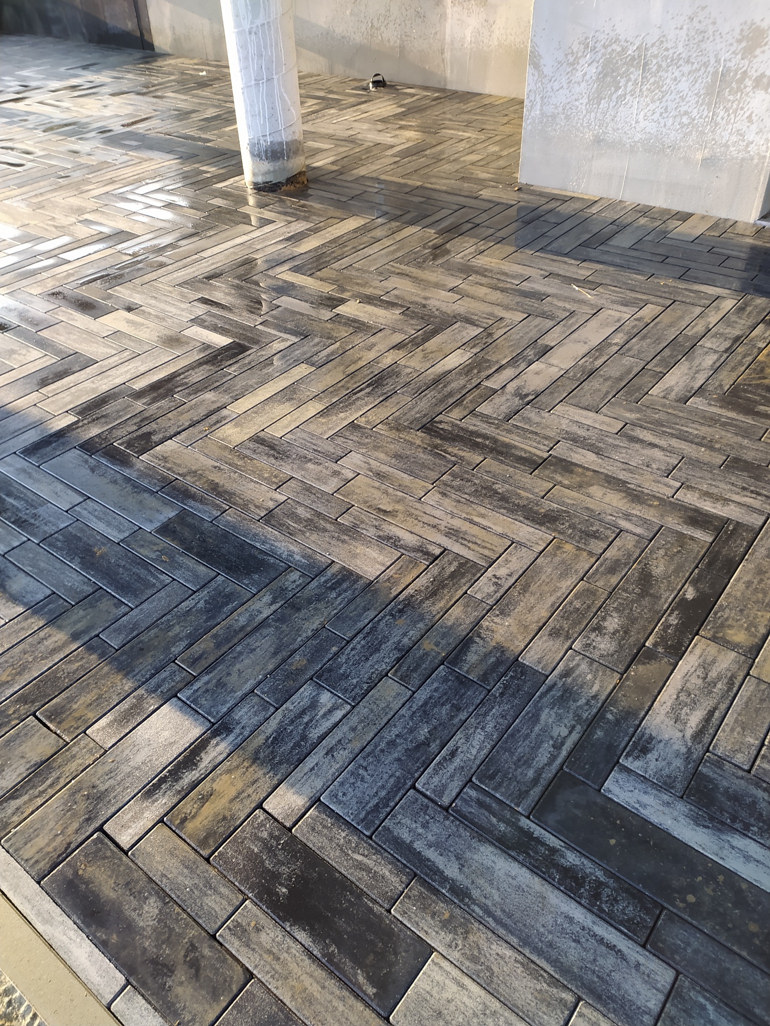 Ułożona kostka brukowa w wzór jodełki, mokra od deszczu, z widocznym słupem zabezpieczonym folią i fragmentem ściany w tle.