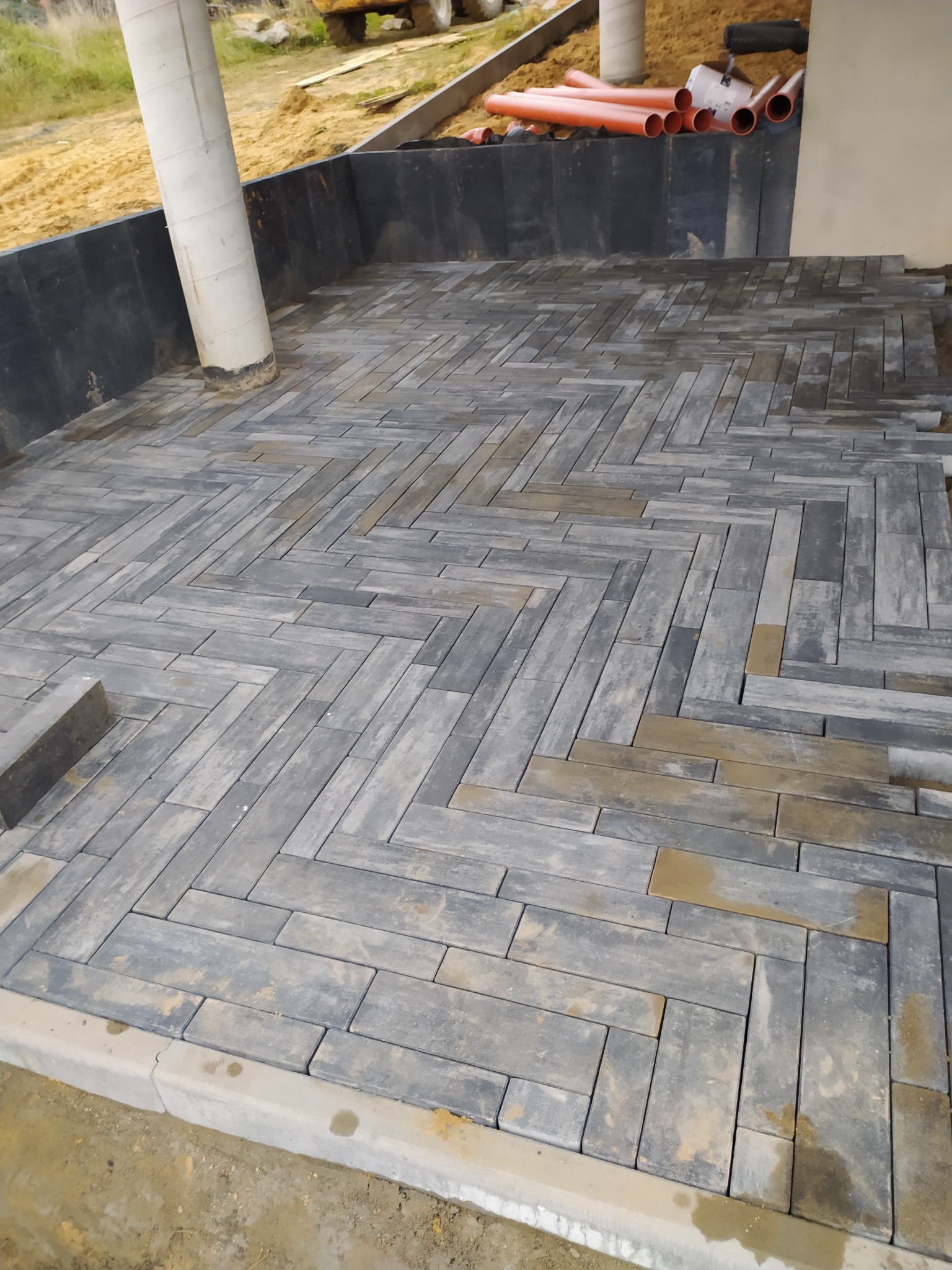 Świeżo ułożona, szara kostka brukowa w jodełkę na posesji w Kolbuszowej, z widocznymi elementami przygotowania terenu i rurami w tle.