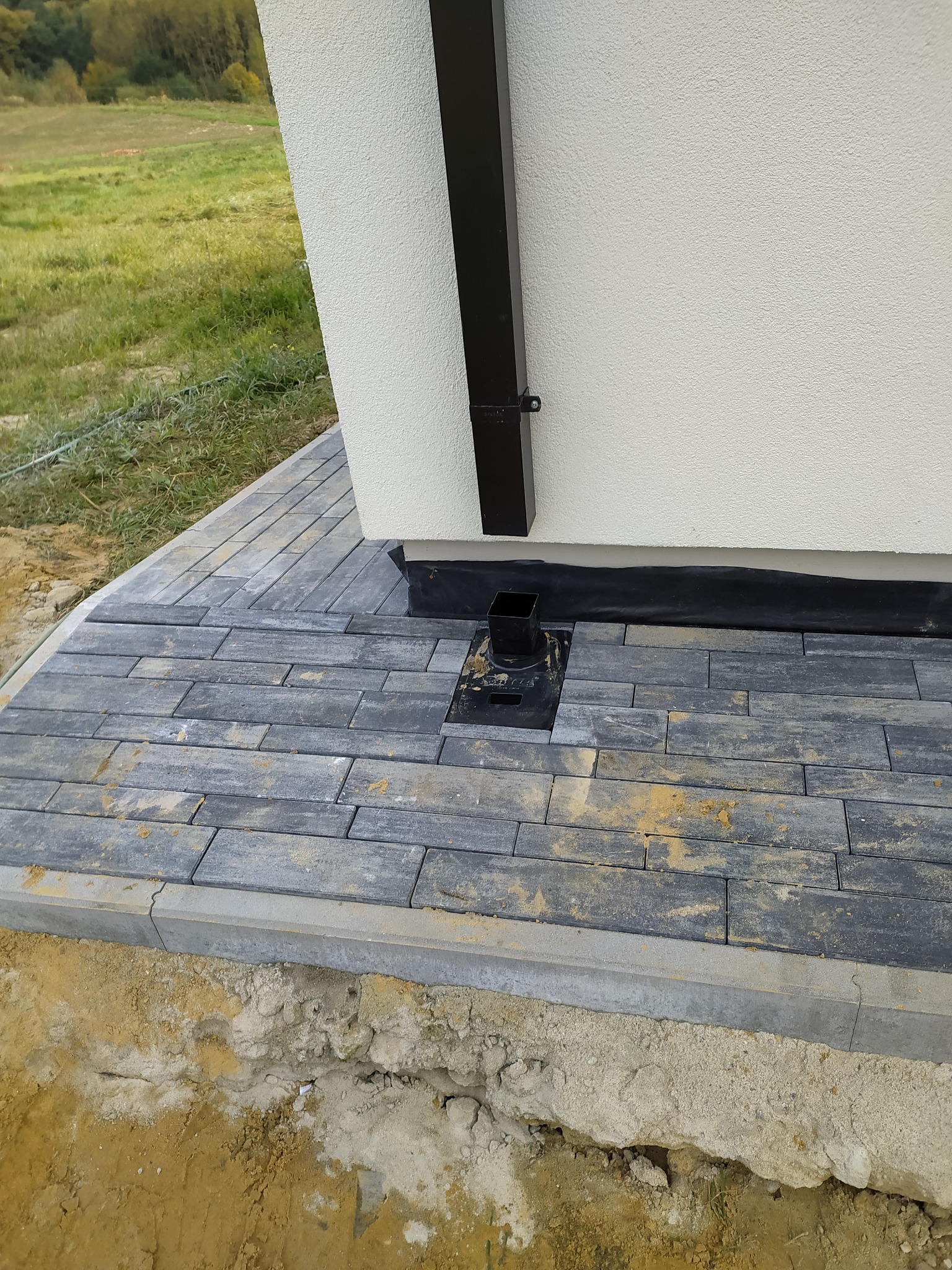 Ułożona, szara kostka brukowa przy ścianie budynku z rynną spustową, widoczna studzienka odpływowa i obrzeże kostki z odsłoniętą ziemią.