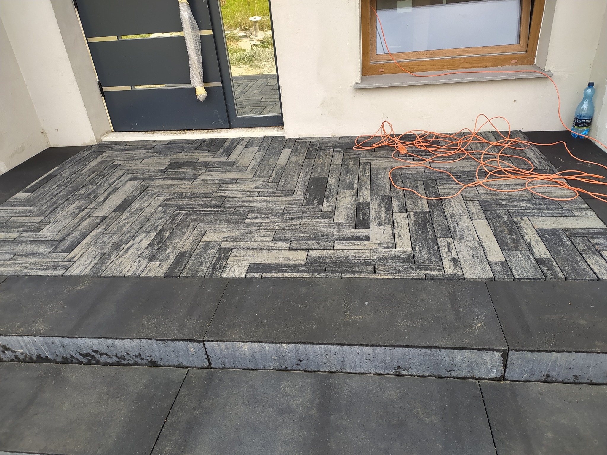 Ułożona kostka brukowa w jodełkę przed wejściem do domu, widoczne przewody elektryczne i butelka z wodą przy oknie.