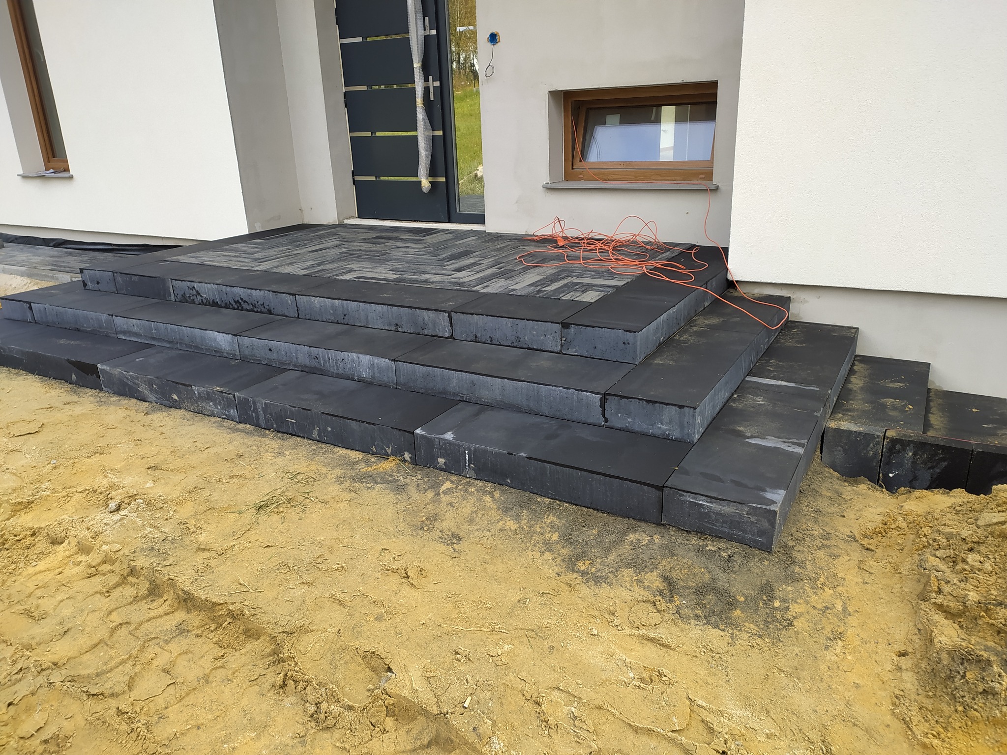 Schody wejściowe wyłożone ciemną kostką brukową w trakcie montażu, widoczny kabel elektryczny i odsłonięta ziemia.