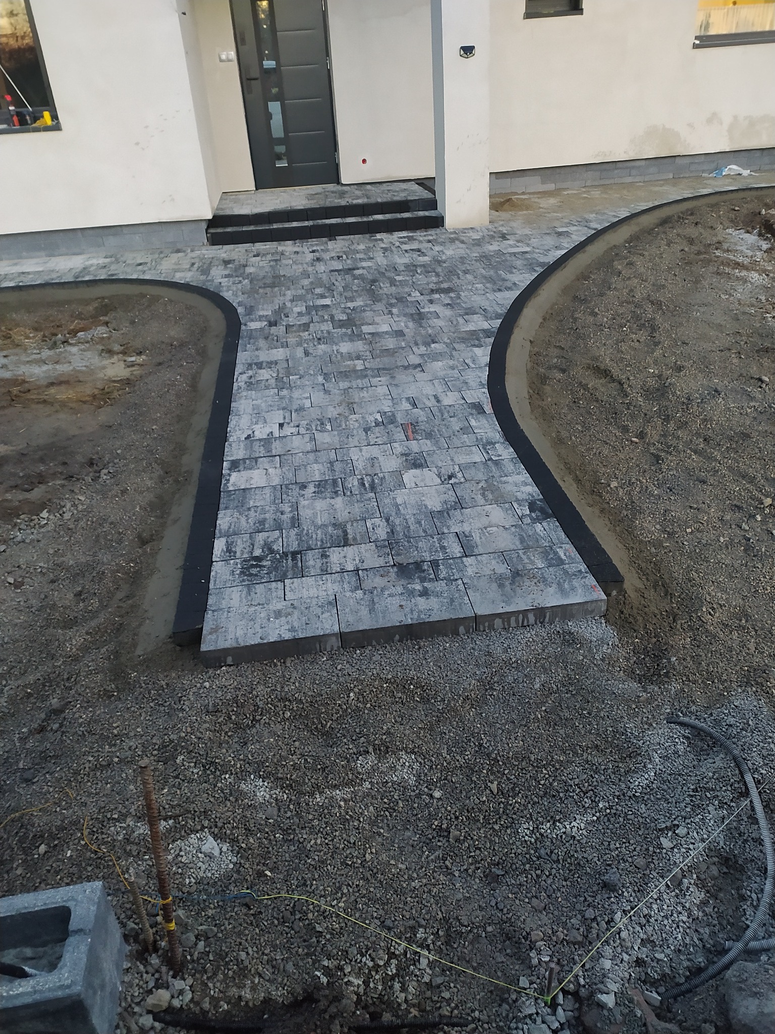 Ścieżka z szarej kostki brukowej prowadząca do wejścia domu, obrzeża wykończone czarnym elementem, teren wokół w trakcie przygotowania pod dalsze prace.