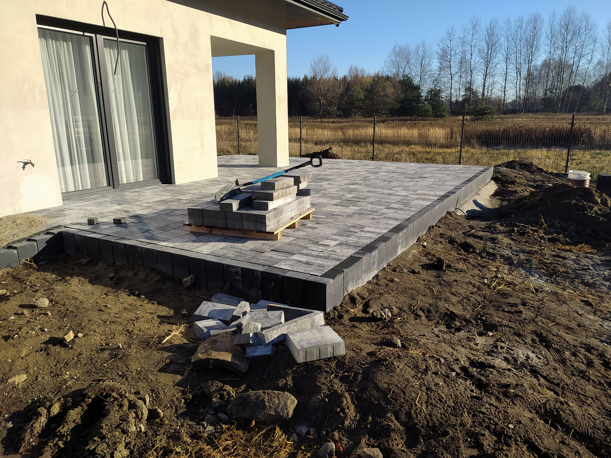 Fragment tarasu z kostki brukowej w trakcie budowy, widoczny stos kostki na palecie, łopata i ziemia wokół, na tle domu i drzew