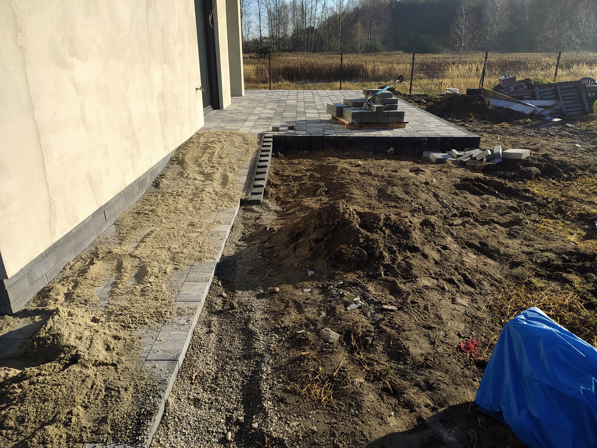 Fragment posesji w trakcie prac brukarskich: ułożona część ścieżki z kostki, przygotowane podłoże z piasku, paleta z kostką oraz pryzma ziemi z nieuprzątniętymi resztkami po budowie.