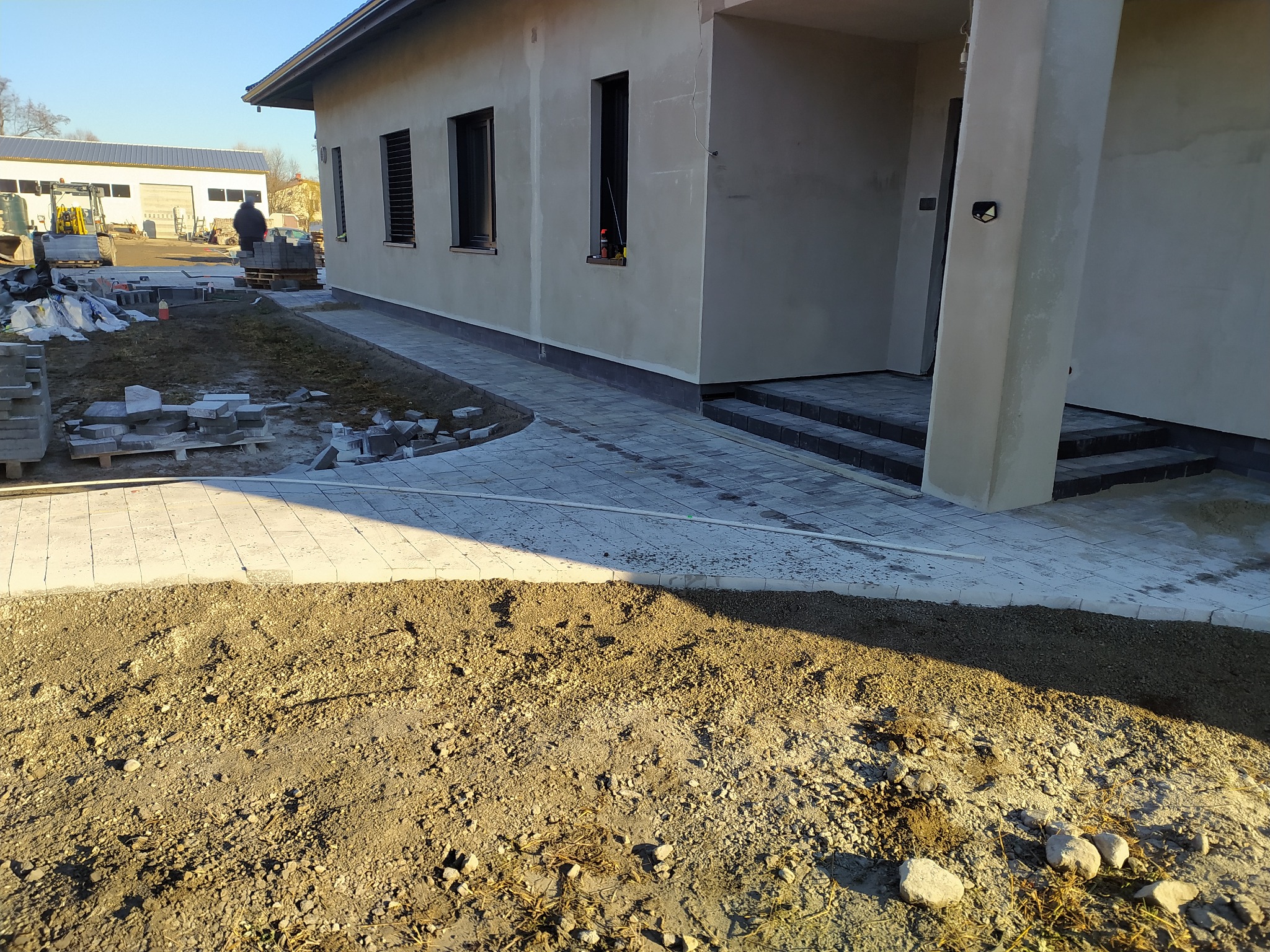 Świeżo ułożona jasnoszara kostka brukowa przed domem w trakcie wykańczania, widoczne palety z kostką, ziemia przygotowana pod dalsze prace, budynek w stanie surowym.