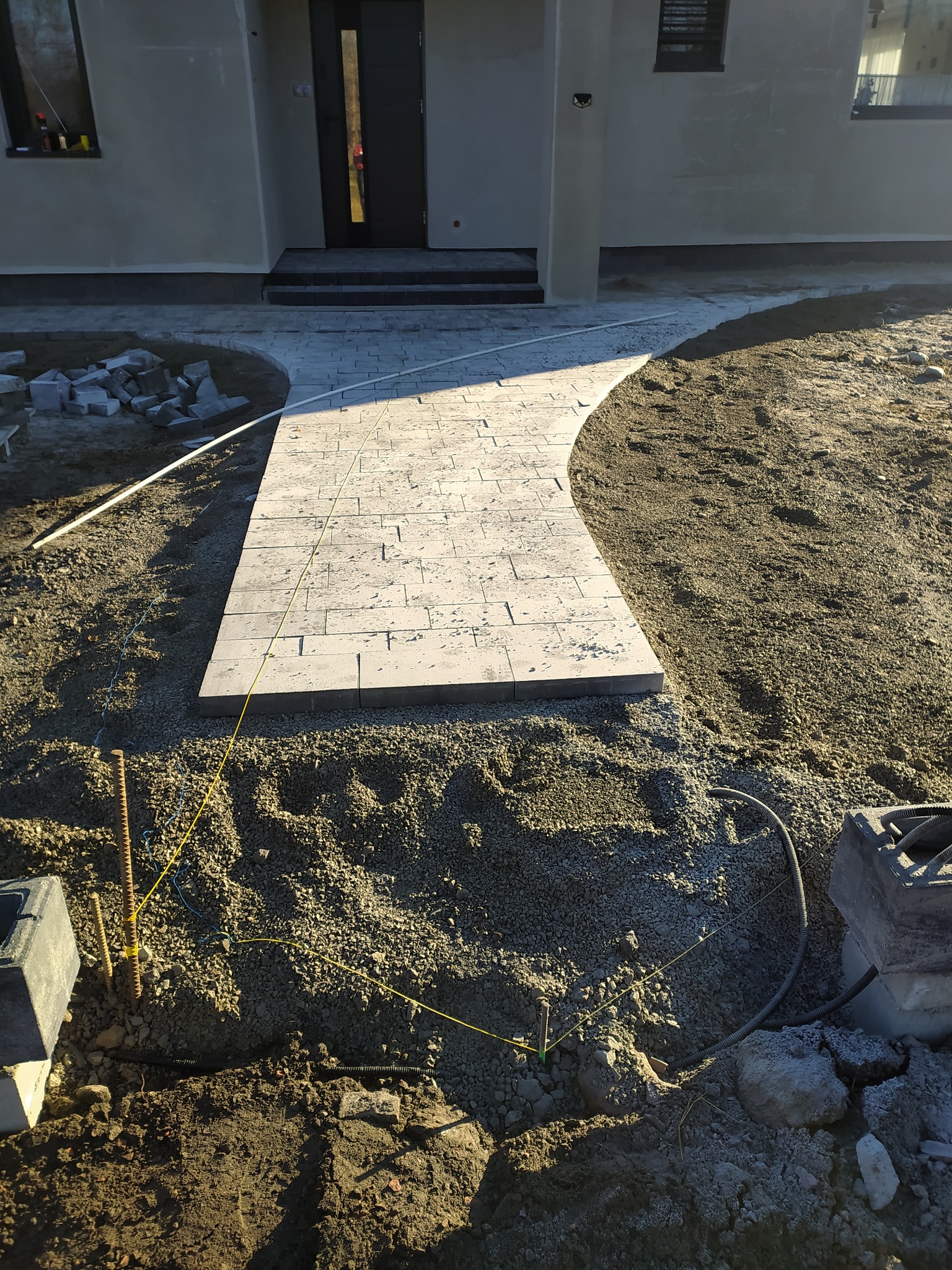 Świeżo ułożona ścieżka z jasnoszarej kostki brukowej prowadząca do drzwi wejściowych domu, z widocznymi elementami przygotowania terenu i niezakończonymi pracami wykończeniowymi wokół.
