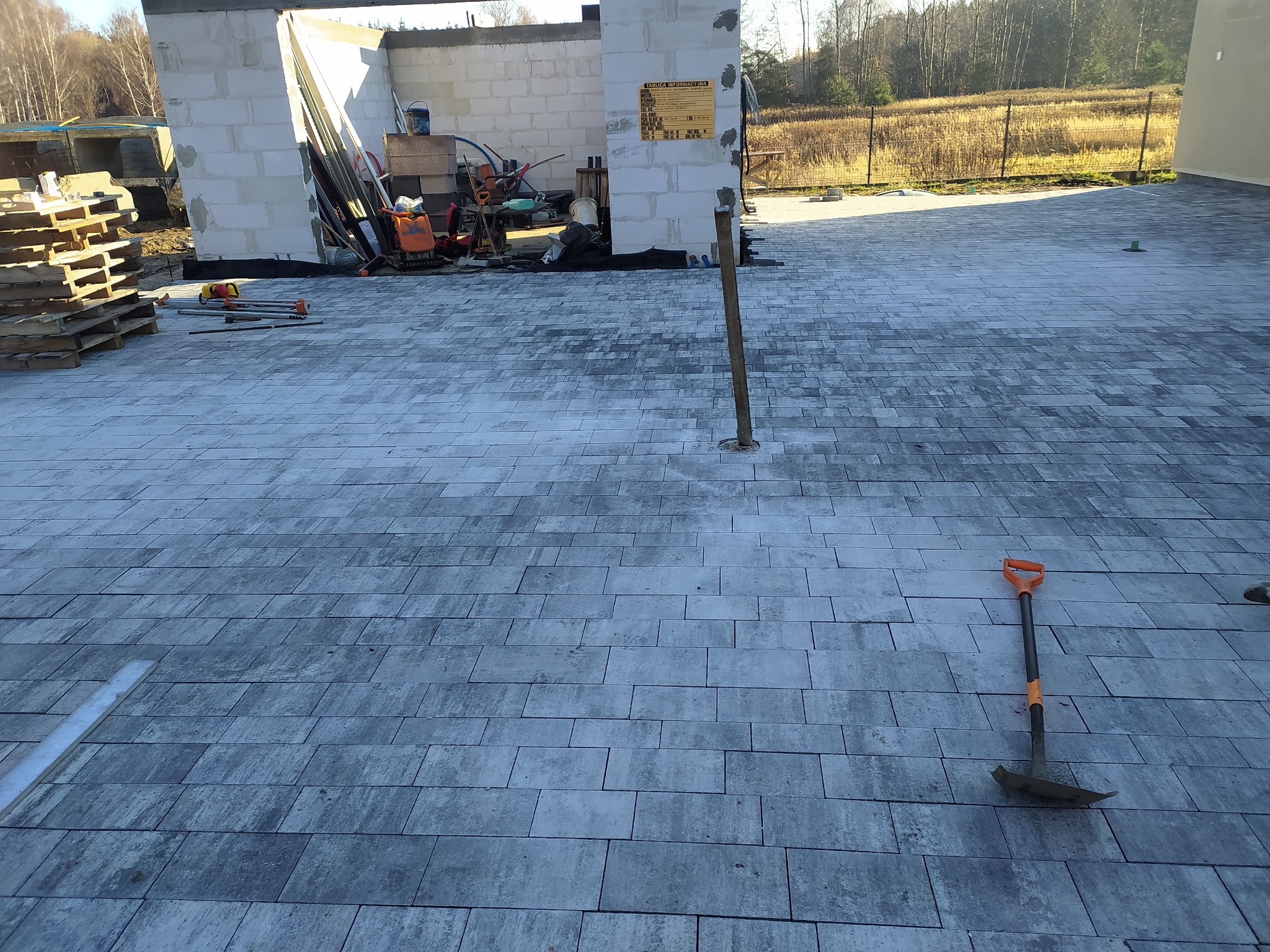 Ułożona kostka brukowa w szarych odcieniach na placu budowy z widoczną łopatą i paletami, w tle surowy budynek w trakcie wznoszenia.