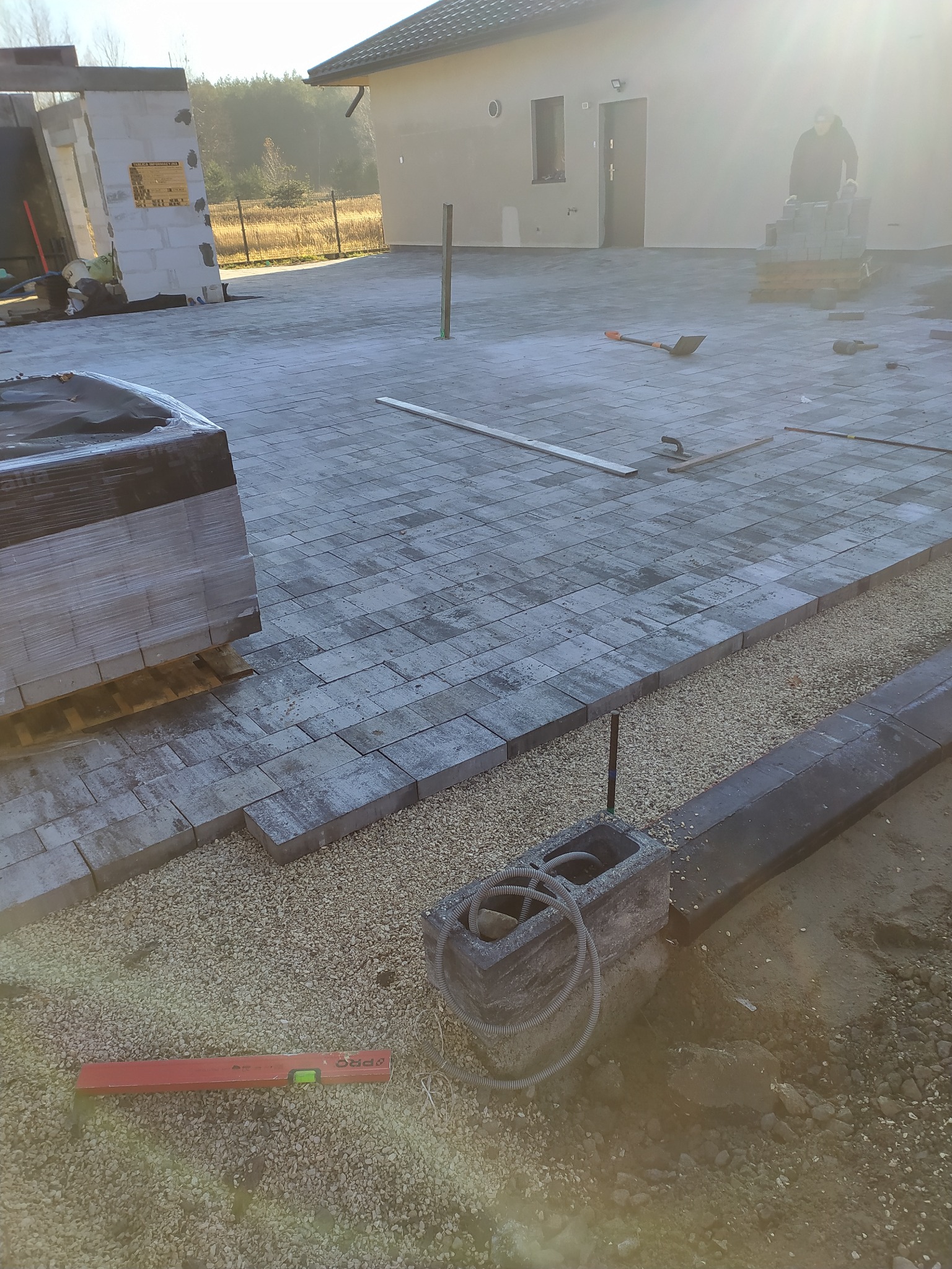 Fragment układanej nawierzchni z kostki brukowej, widoczne podsypka żwirowa, paleta z kostką w folii, poziomica i pracownik w tle.