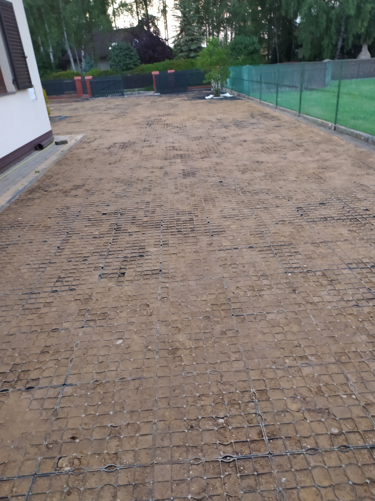 Przygotowany teren pod kostkę brukową z rozłożoną siatką wzmacniającą, widok z boku domu, w tle ogrodzenie i zieleń.