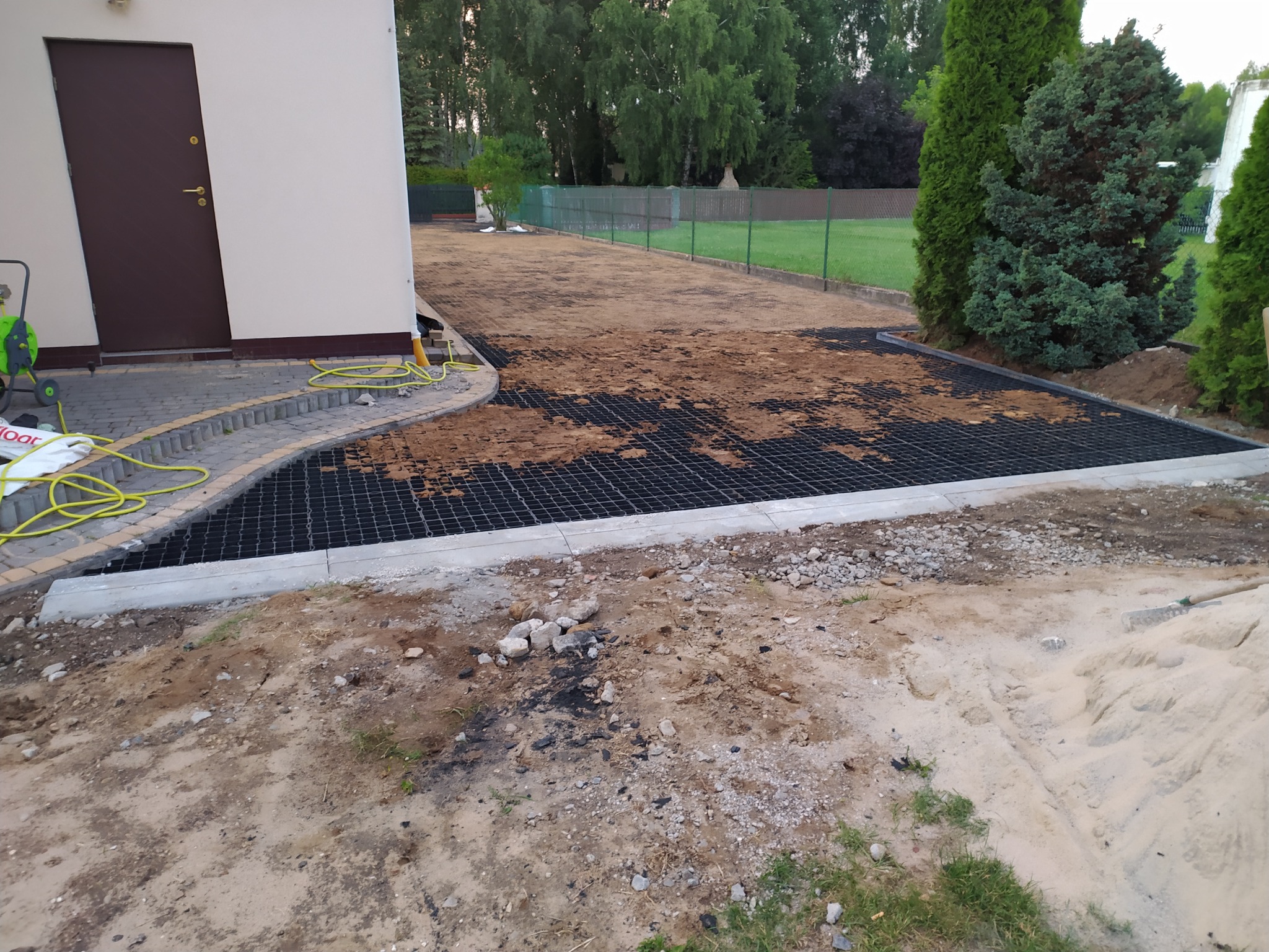 Przygotowanie podłoża pod nawierzchnię: widoczne obrzeża betonowe, geokrata wypełniana ziemią, częściowo zagęszczona, w tle trawnik i budynek z brązowymi drzwiami.