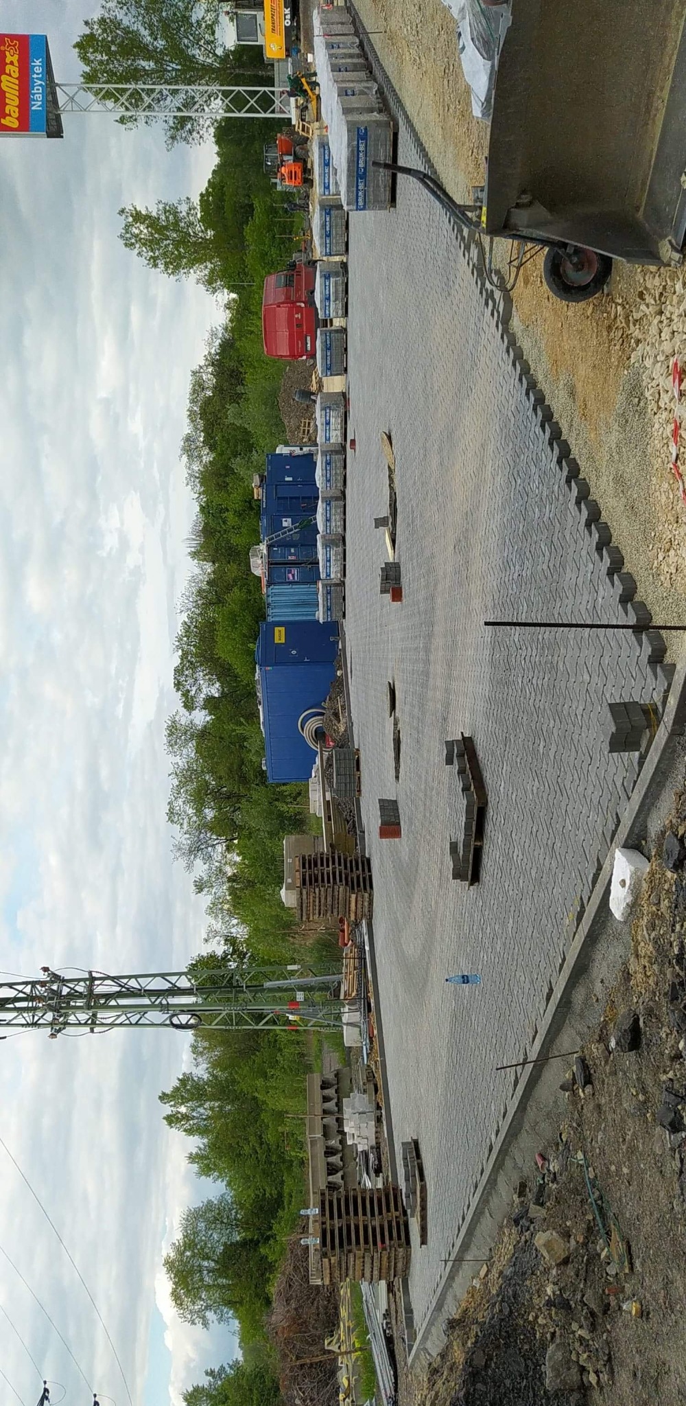 Układanie szarej kostki brukowej na dużej powierzchni, widoczne palety z kostką, maszyny budowlane w tle, teren budowy.