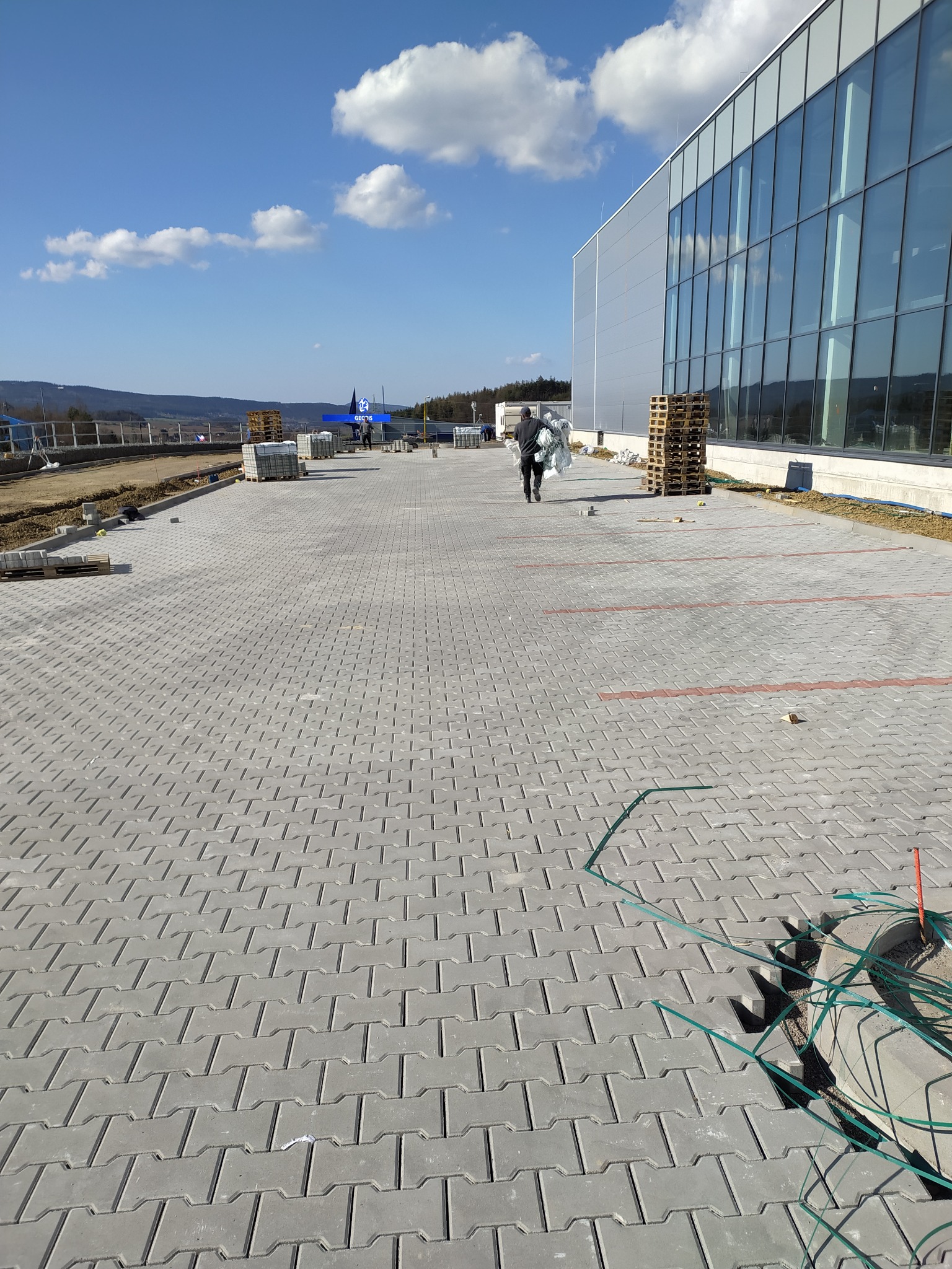 Nowa, szara kostka brukowa ułożona na parkingu przed budynkiem z przeszkloną fasadą; w tle widoczny pracownik z pakietem materiałów, palety z kostką i błękitne niebo z chmurami.