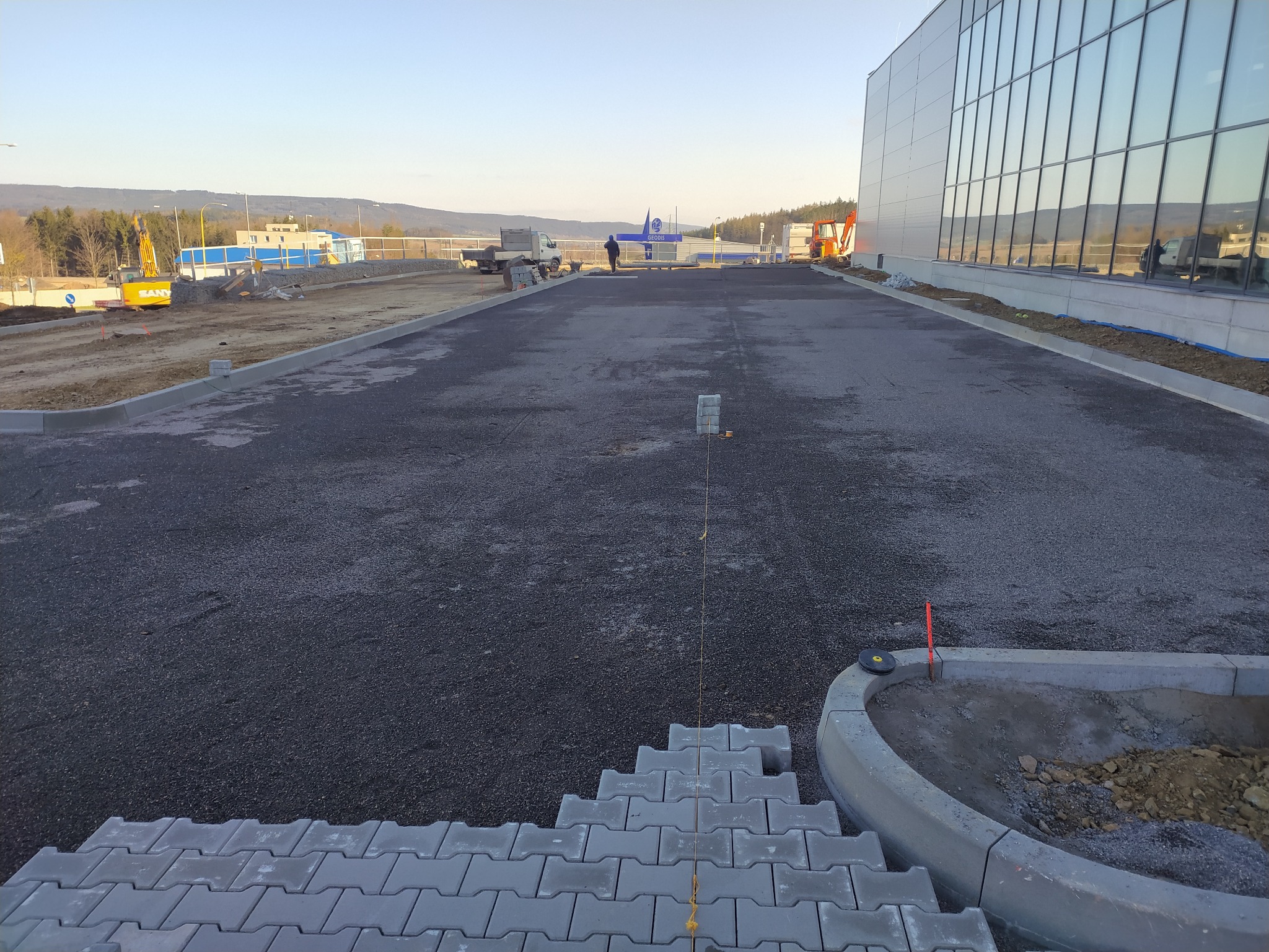 Widok z góry na plac budowy z częściowo ułożoną szarą kostką brukową, w tle widoczny budynek z dużymi oknami, maszyny budowlane i krajobraz z górami.