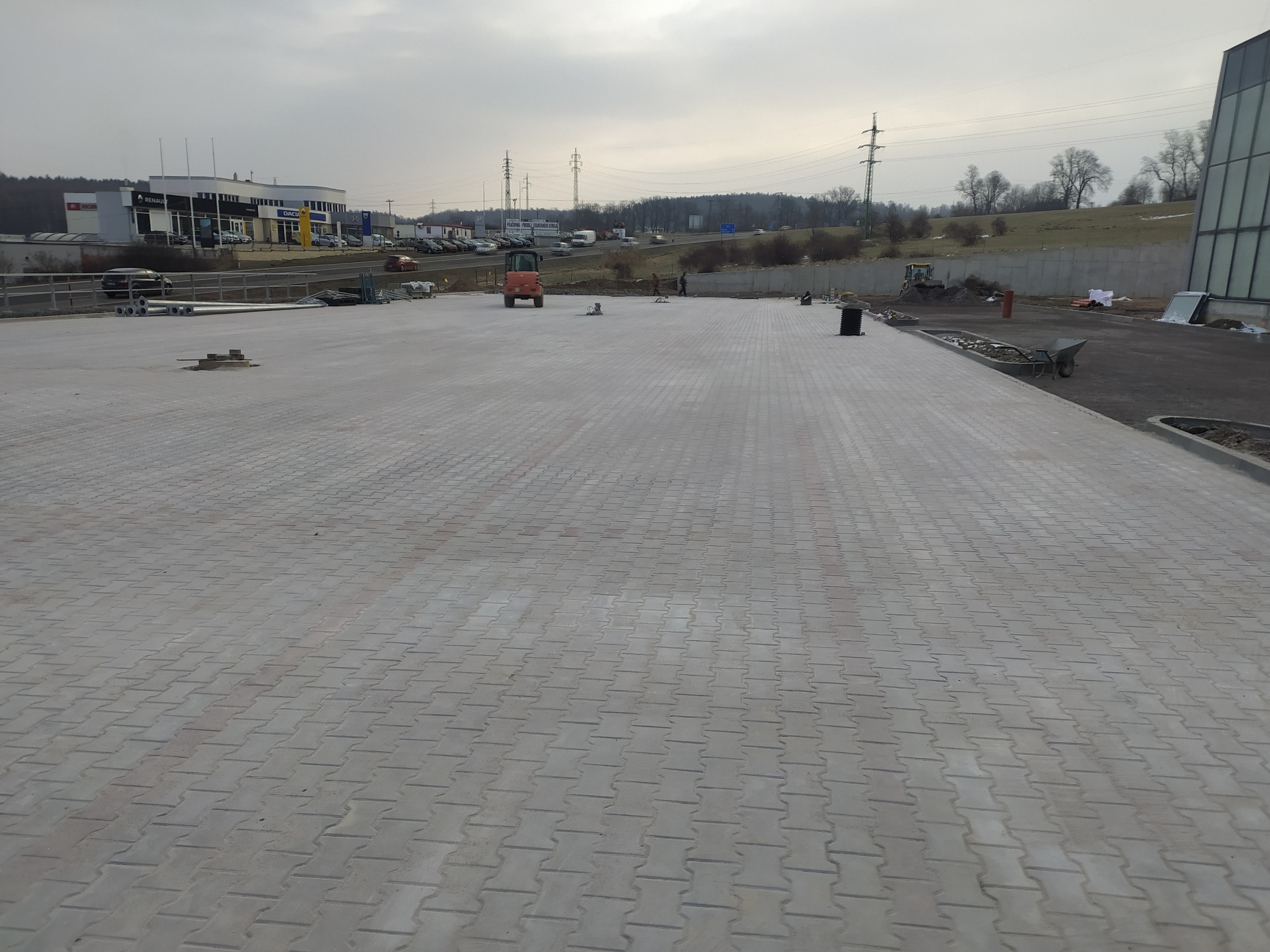 Rozległa, nowo ułożona szara kostka brukowa na terenie inwestycyjnym, z widocznym walcem drogowym w oddali i budynkami handlowymi w tle, pod pochmurnym niebem.