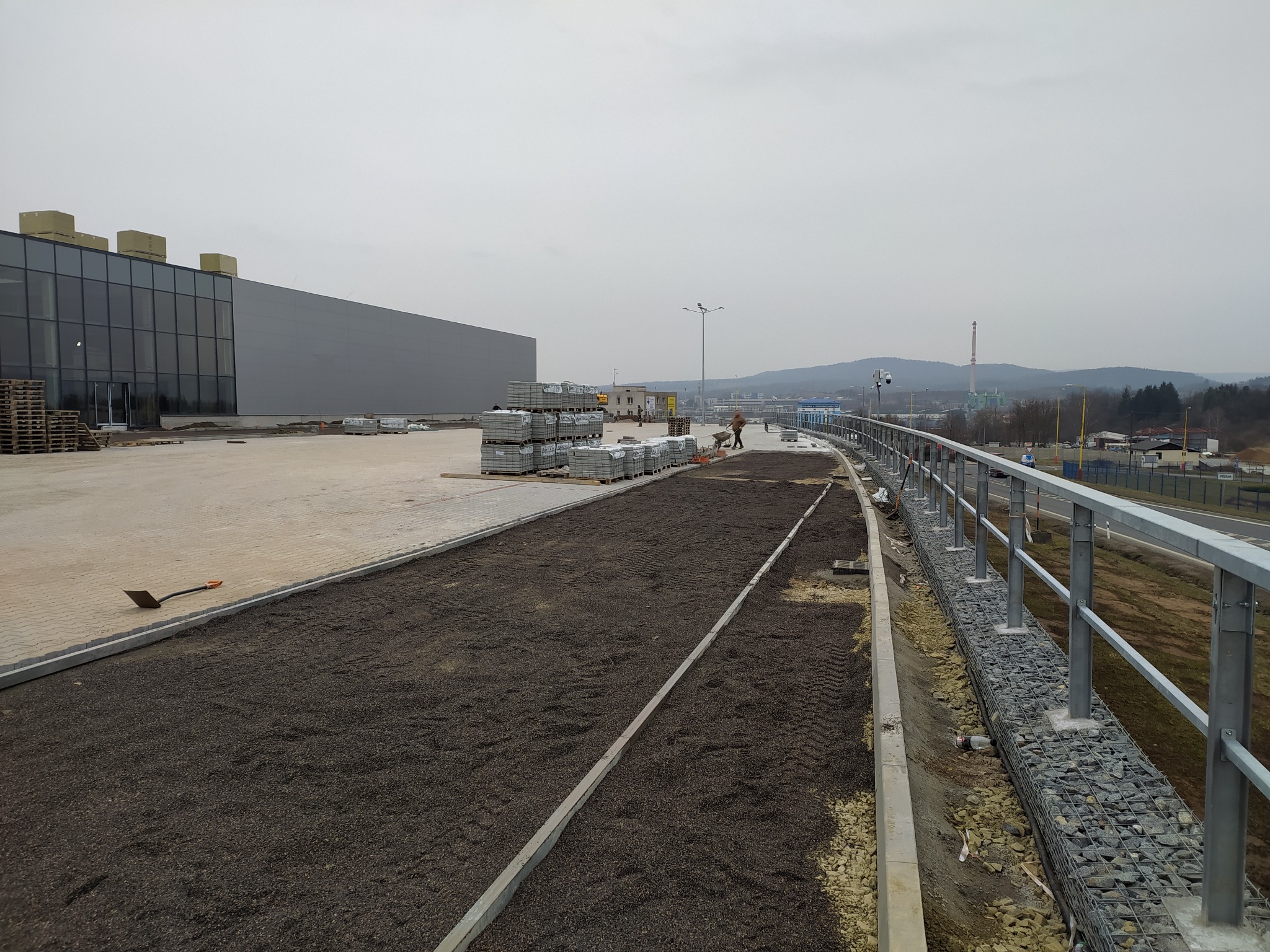 Przygotowanie podłoża pod kostkę brukową na dużej powierzchni, widoczne ułożone krawężniki, podsypka, palety z kostką i pracownik z taczką w oddali.