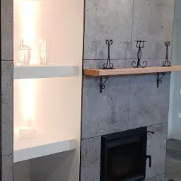 Nowoczesny kominek obłożony szarymi płytami, z drewnianą półką i dekoracyjnymi świecznikami, obok wnęka z oświetlonymi półkami, na których stoją szklane ozdoby i doniczki z roślinami.