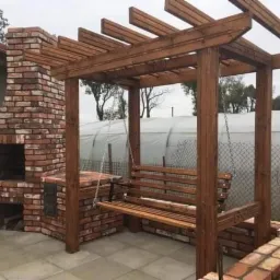 Zewnętrzny kącik wypoczynkowy z murowanym grillem z cegły i drewnianą konstrukcją z huśtawką, na tle szklarni i ogrodzenia z siatki.