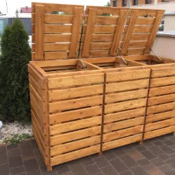 Drewniana obudowa na trzy pojemniki na śmieci z otwartymi klapami, widoczna na zewnątrz budynku, obok zielonego pojemnika i ozdobnego krzewu.