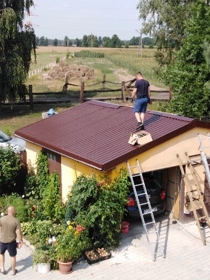 Prace dekarskie na dachu garażu z blachy trapezowej w kolorze brązowym, widok z góry na pracownika z pudełkiem narzędzi, drabiny opierają się o budynek, w tle wiejski krajobraz.