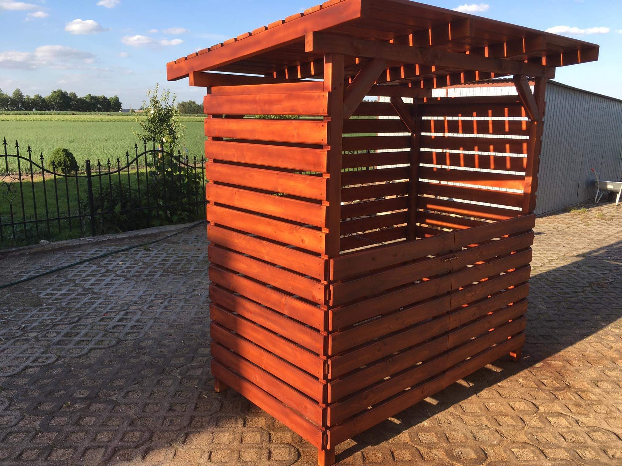 Drewniana konstrukcja z poziomych listew w kolorze ciepłego brązu, prawdopodobnie altana, ustawiona na betonowej kostce brukowej, z widokiem na zielone pole i ogrodzenie w tle.