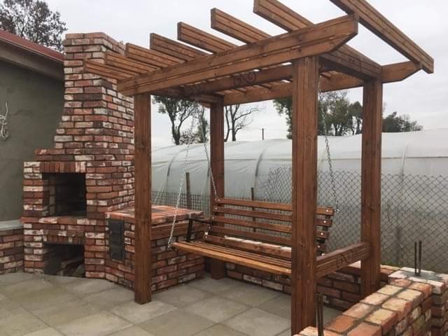 Zewnętrzny kącik wypoczynkowy z murowanym grillem z cegły i drewnianą konstrukcją z huśtawką, na tle szklarni i ogrodzenia z siatki.