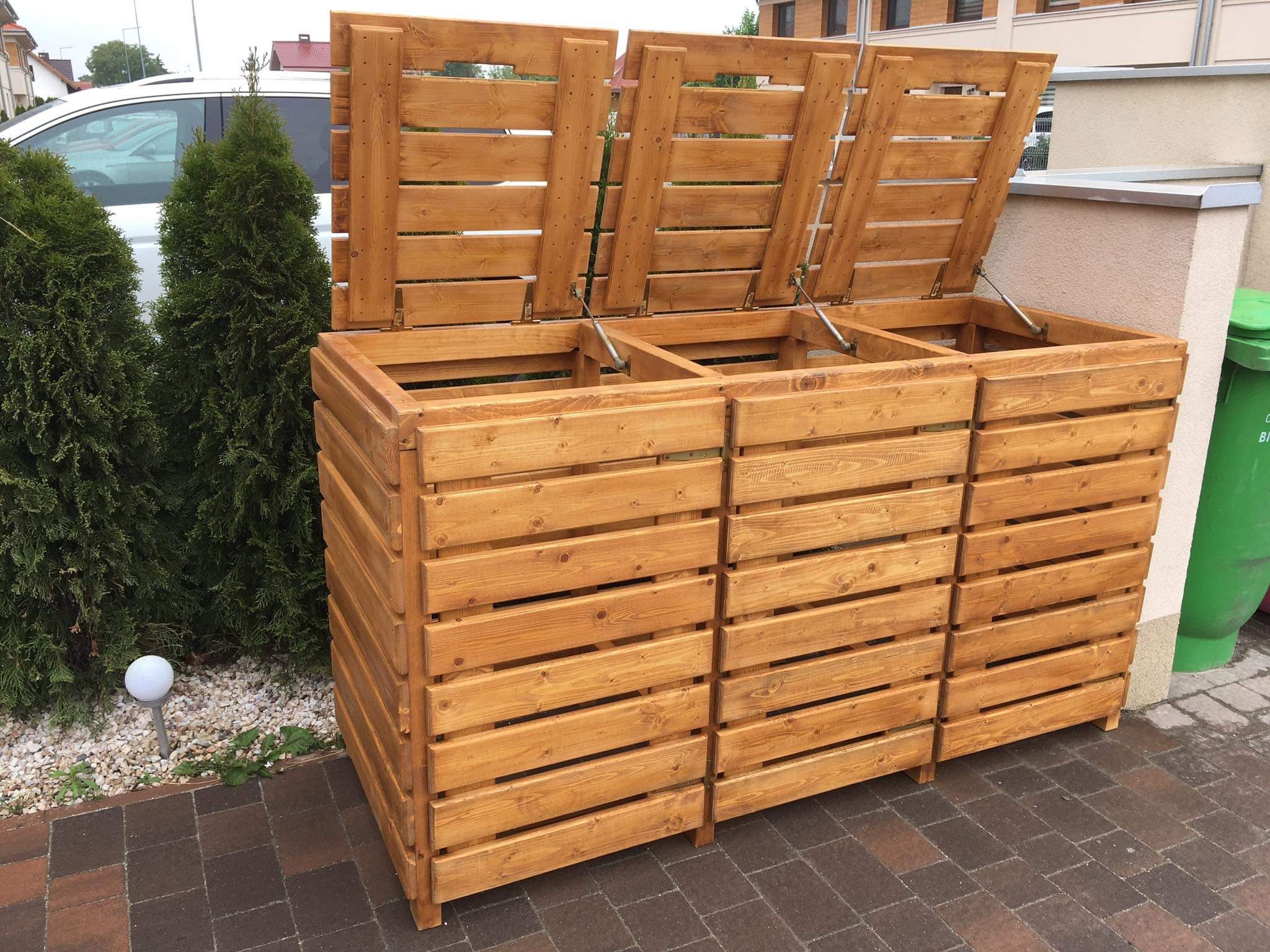 Drewniana obudowa na trzy pojemniki na śmieci z otwartymi klapami, widoczna na zewnątrz budynku, obok zielonego pojemnika i ozdobnego krzewu.