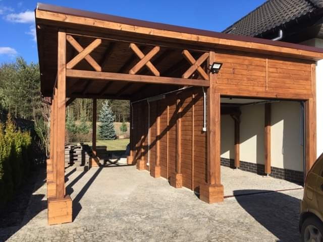 Drewniana konstrukcja zadaszenia nad podjazdem, z dekoracyjnymi krzyżowymi belkami, połączona z garażem w domu jednorodzinnym, oświetlona lampą zewnętrzną.