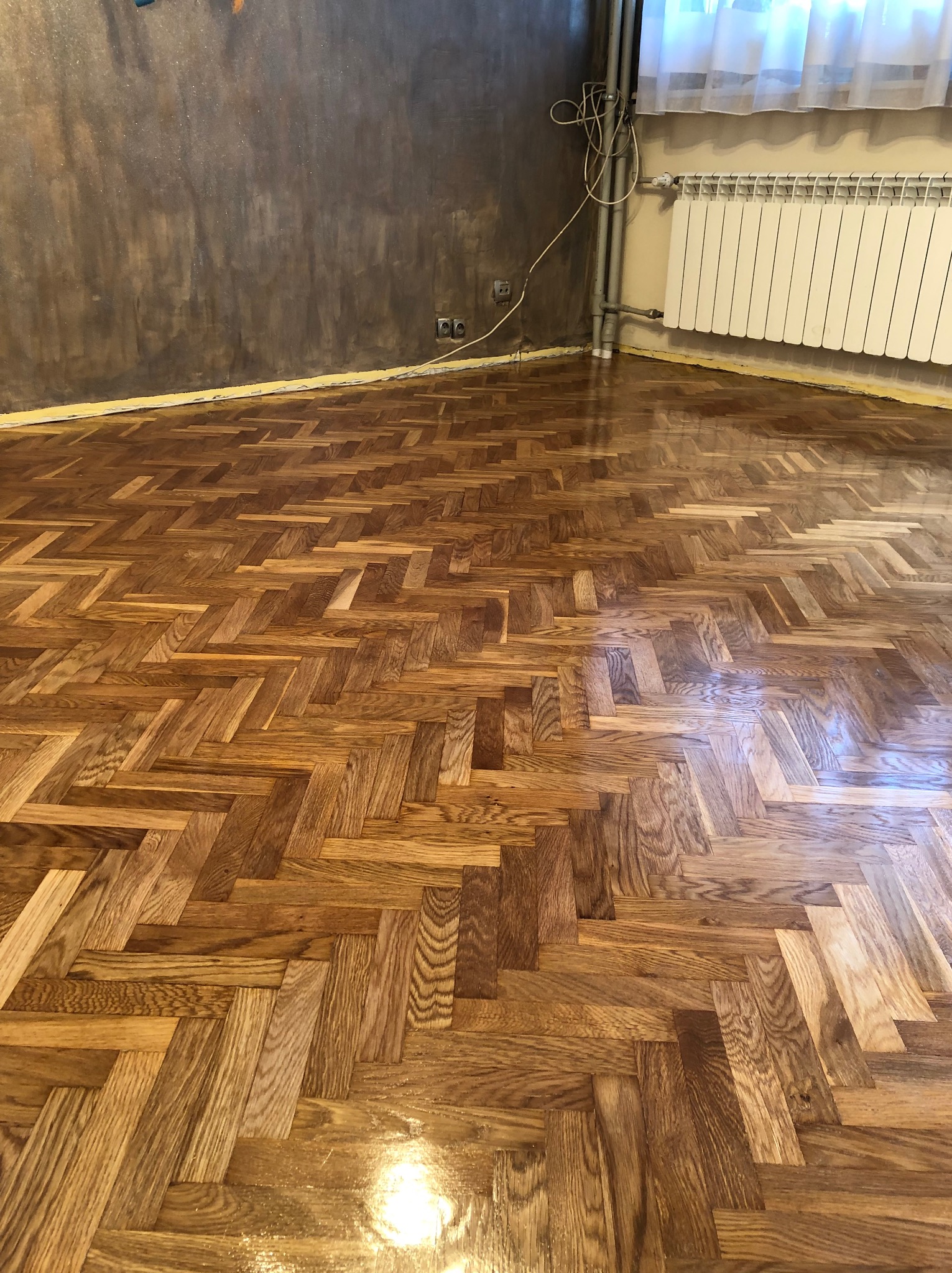 Ułożony parkiet w jodełkę, widoczny fragment szarej ściany i biały grzejnik. Całość pomieszczenia po remoncie, gotowa do użytku.