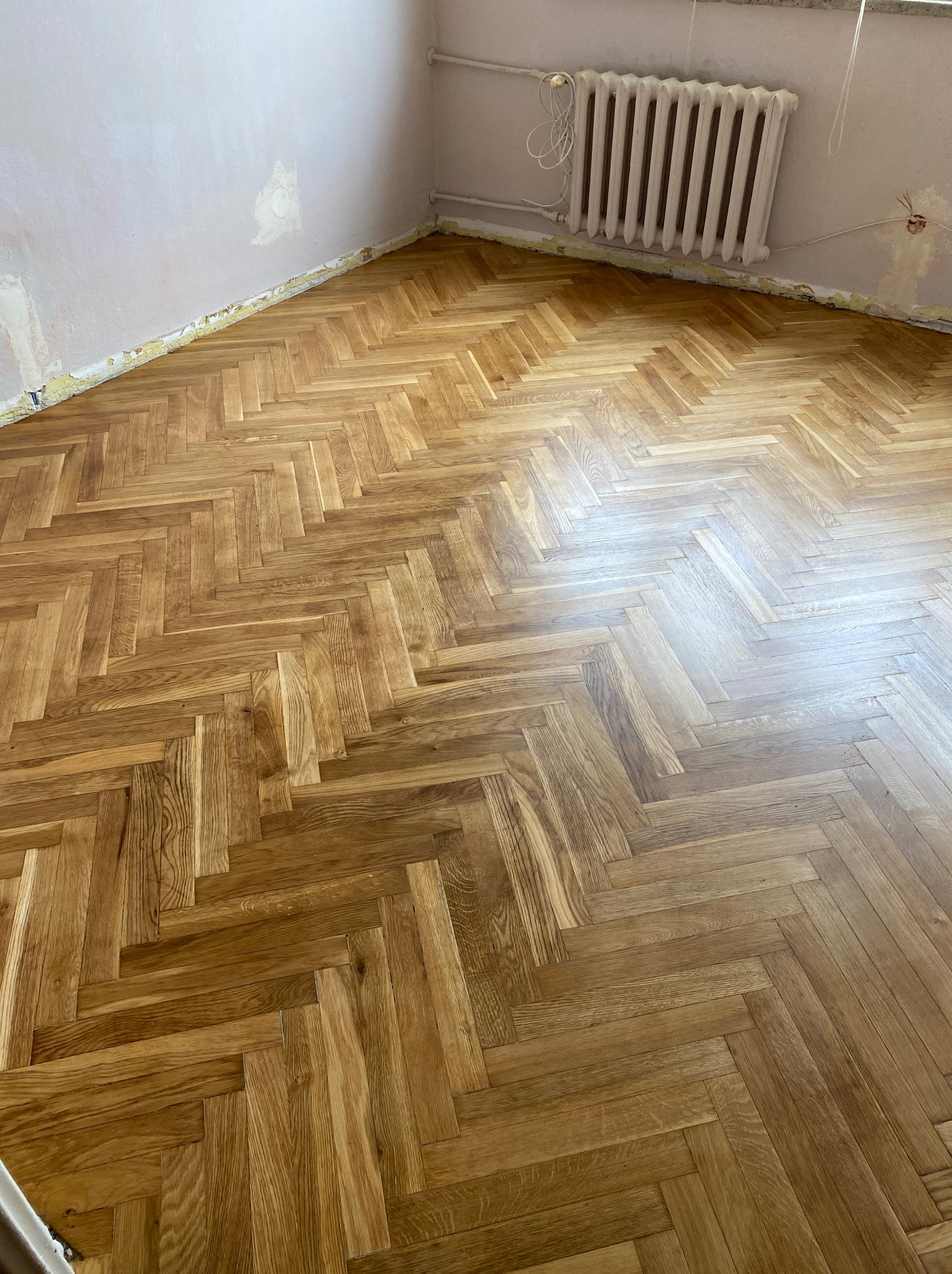 Parkiet ułożony w jodełkę w Warszawskim mieszkaniu. Jasne drewno, widoczny grzejnik i fragment ściany w trakcie przygotowania do malowania. Naturalne światło.