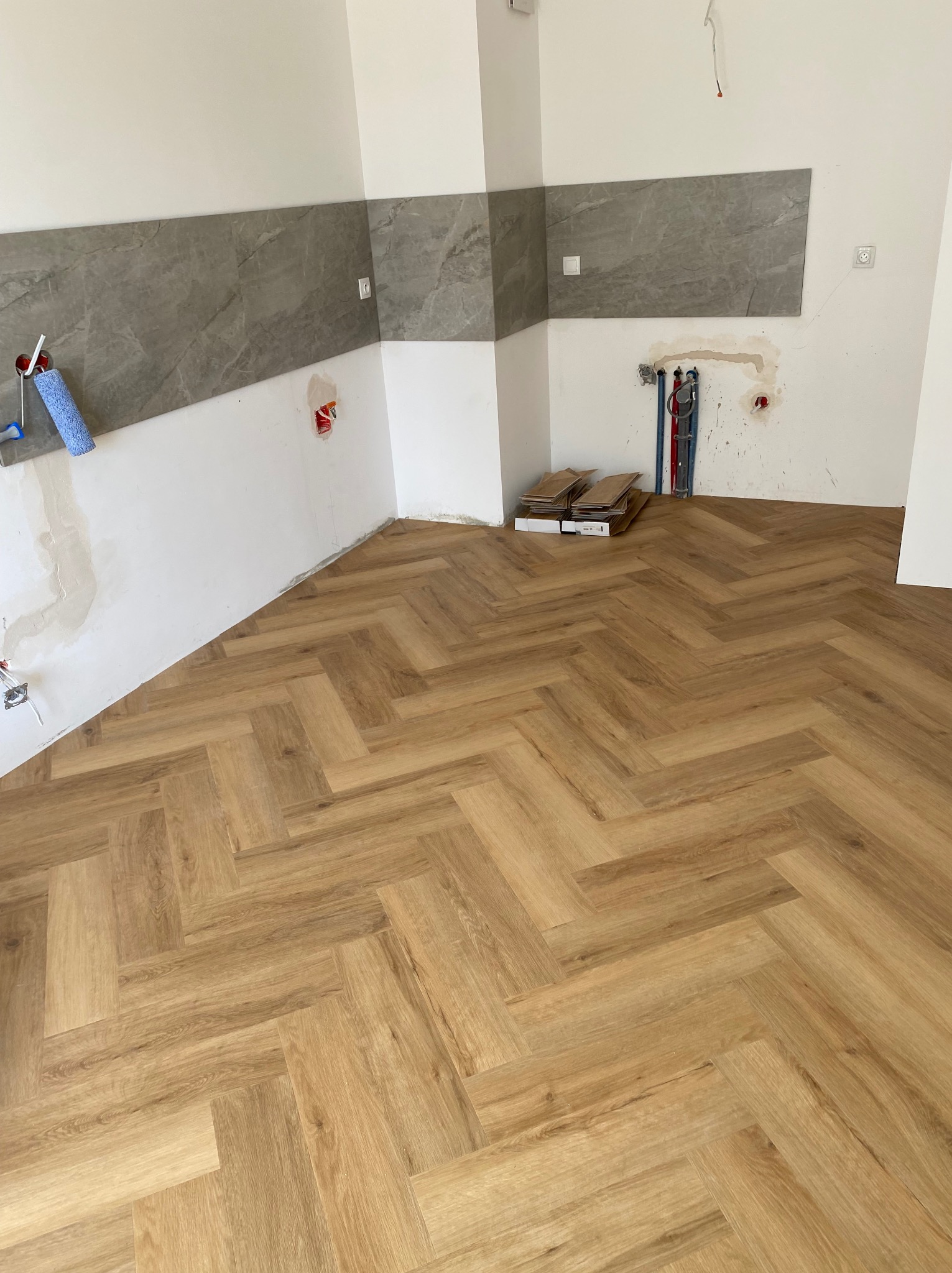 Jasne panele ułożone we wzór jodełki w nowym wnętrzu. Częściowo wykończone ściany z szarym pasem płytek. Widoczne puszki elektryczne i rury hydrauliczne.