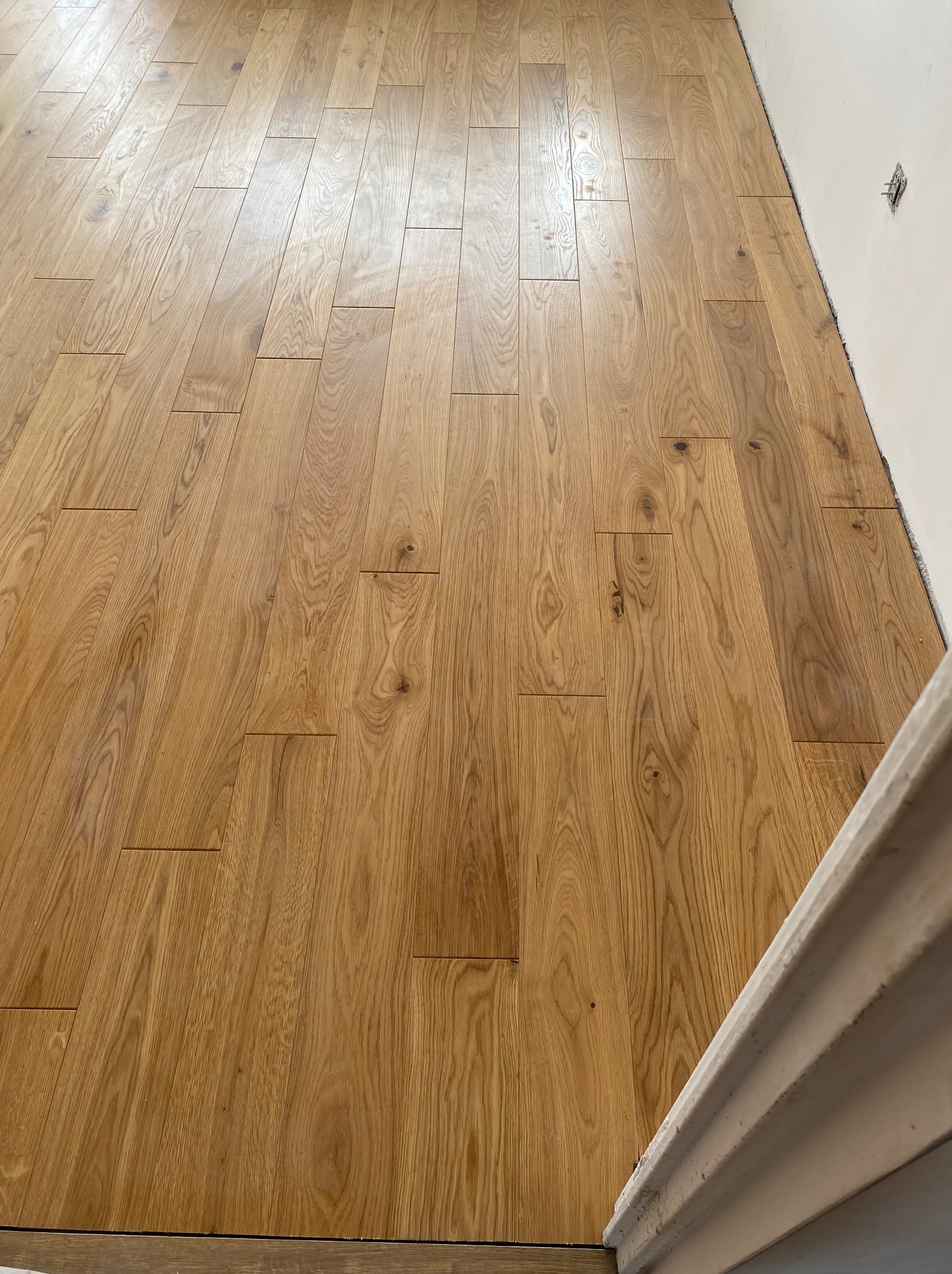 Ułożony parkiet z desek o naturalnym odcieniu drewna, widoczne detale usłojenia. Ściana w tle z wystającą instalacją elektryczną. Ujęcie z niskiego kąta.