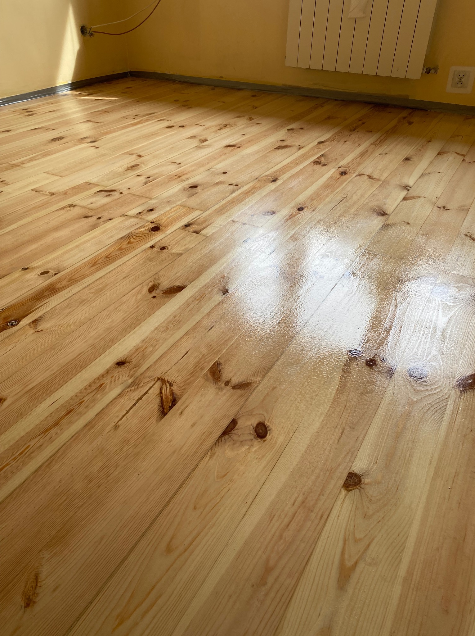 Jasna, lakierowana podłoga z desek sosnowych, widok z góry, z delikatnym połyskiem i widocznymi słojami drewna, przy ścianie grzejnik.