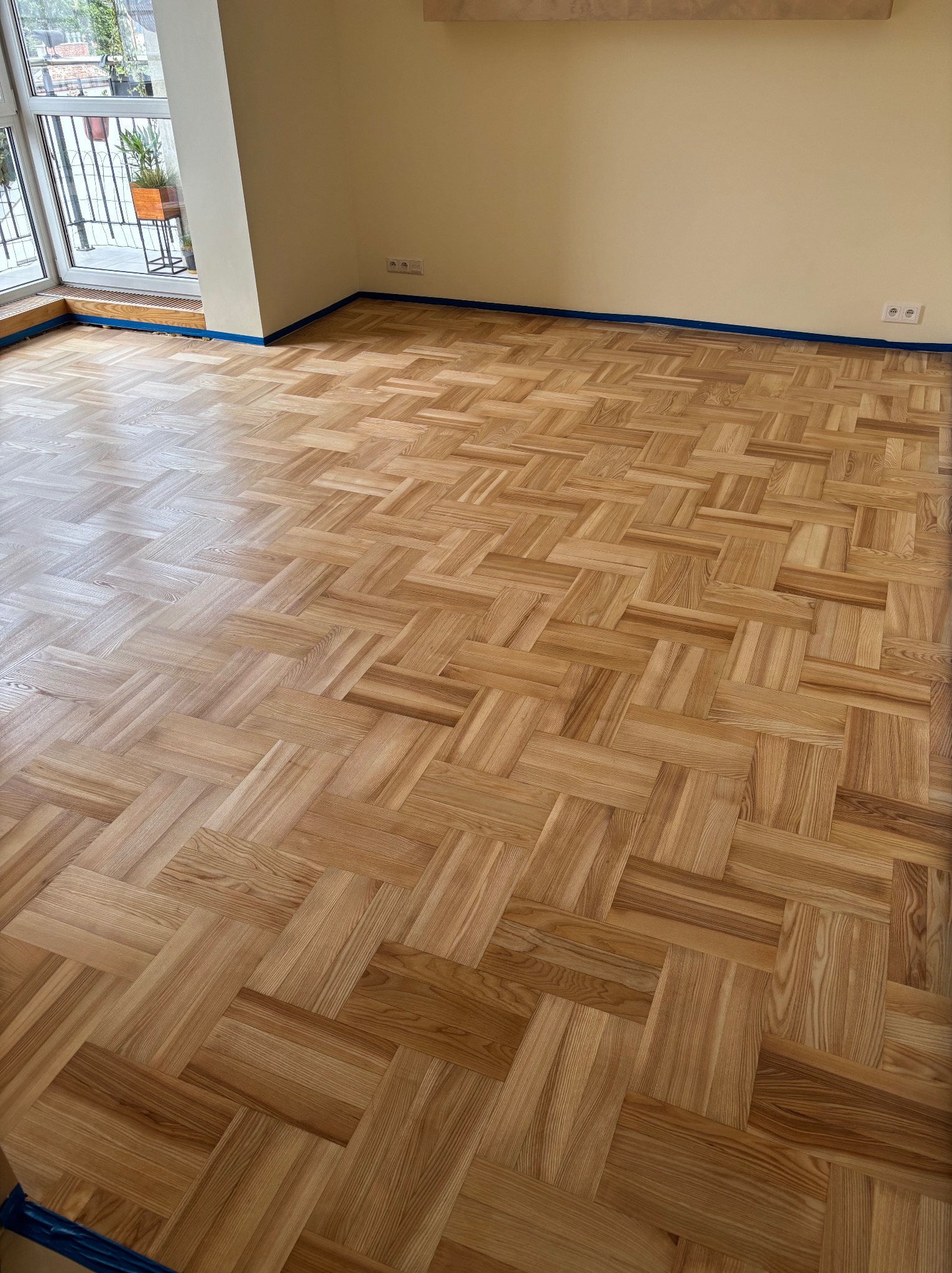 Parkiet ułożony w jodełkę francuską, zabezpieczony niebieską taśmą malarską przy ścianach. Widok na balkon z roślinami w tle. Jasne, naturalne odcienie drewna.