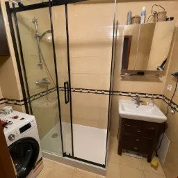 Zainstalowana kabina prysznicowa z czarnymi ramami w łazience z beżowymi płytkami, obok pralka i umywalka z szafką