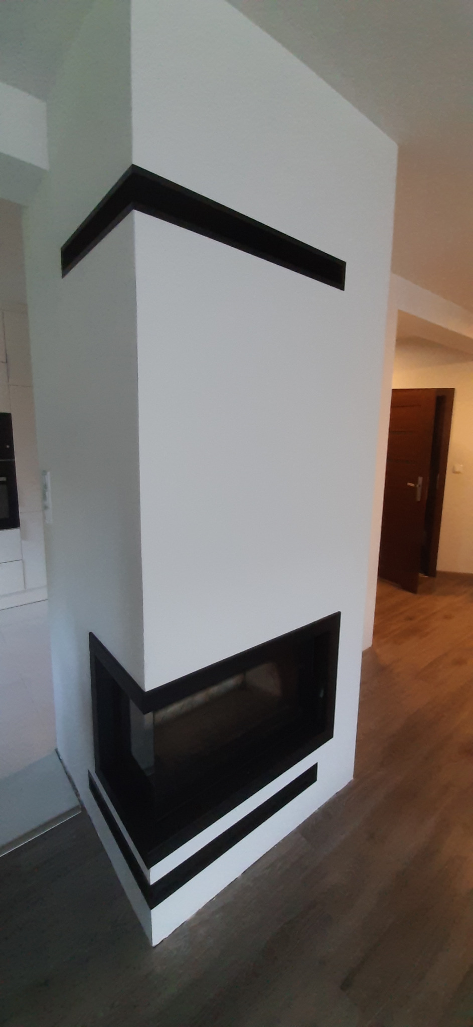 Biały narożny kominek z czarnymi elementami dekoracyjnymi w nowoczesnym salonie z drewnianą podłogą, widok na otwarte drzwi do innego pomieszczenia.