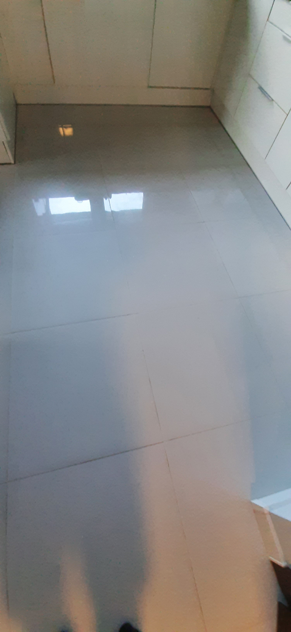 Błyszcząca, jasnoszara podłoga z płytek w kuchni, widoczne odbicie światła sufitowego, białe szafki kuchenne w tle.