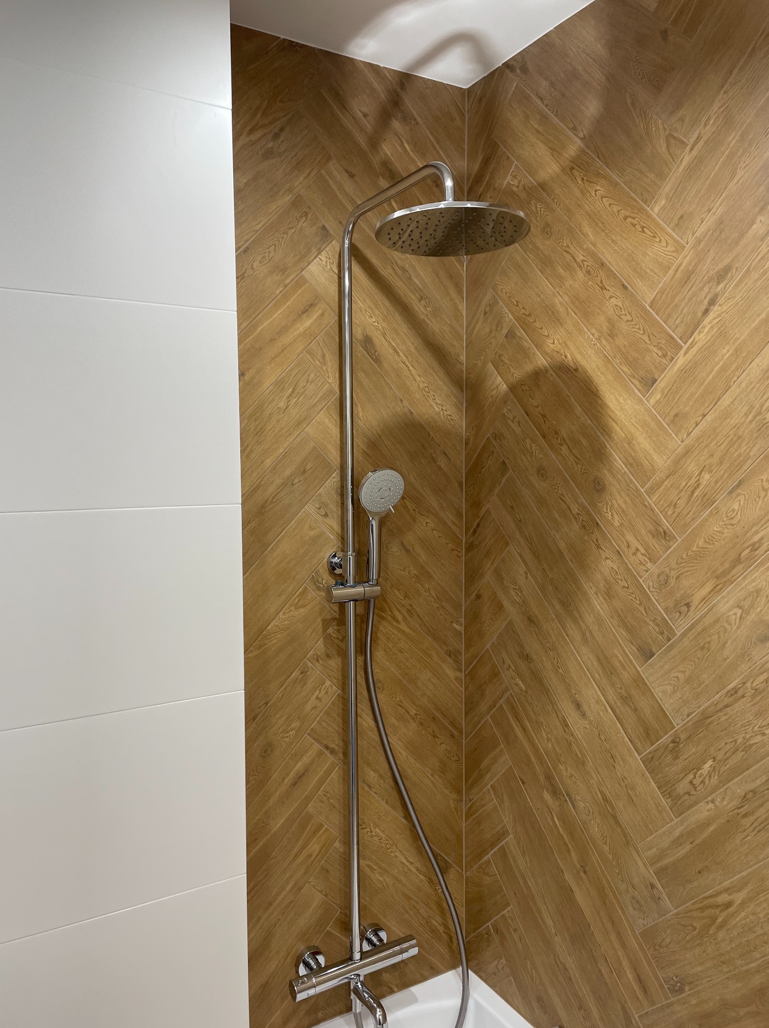 Wykończona kabina prysznicowa z chromowaną deszczownicą i baterią, ściany wyłożone białymi płytkami i płytkami imitującymi drewno ułożonymi w jodełkę.