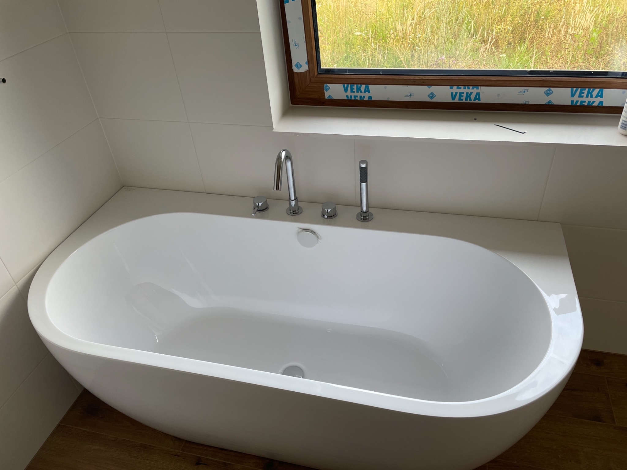Nowoczesna, wolnostojąca wanna owalna z chromowaną armaturą w jasnej łazience z oknem, widok na łąkę za oknem.