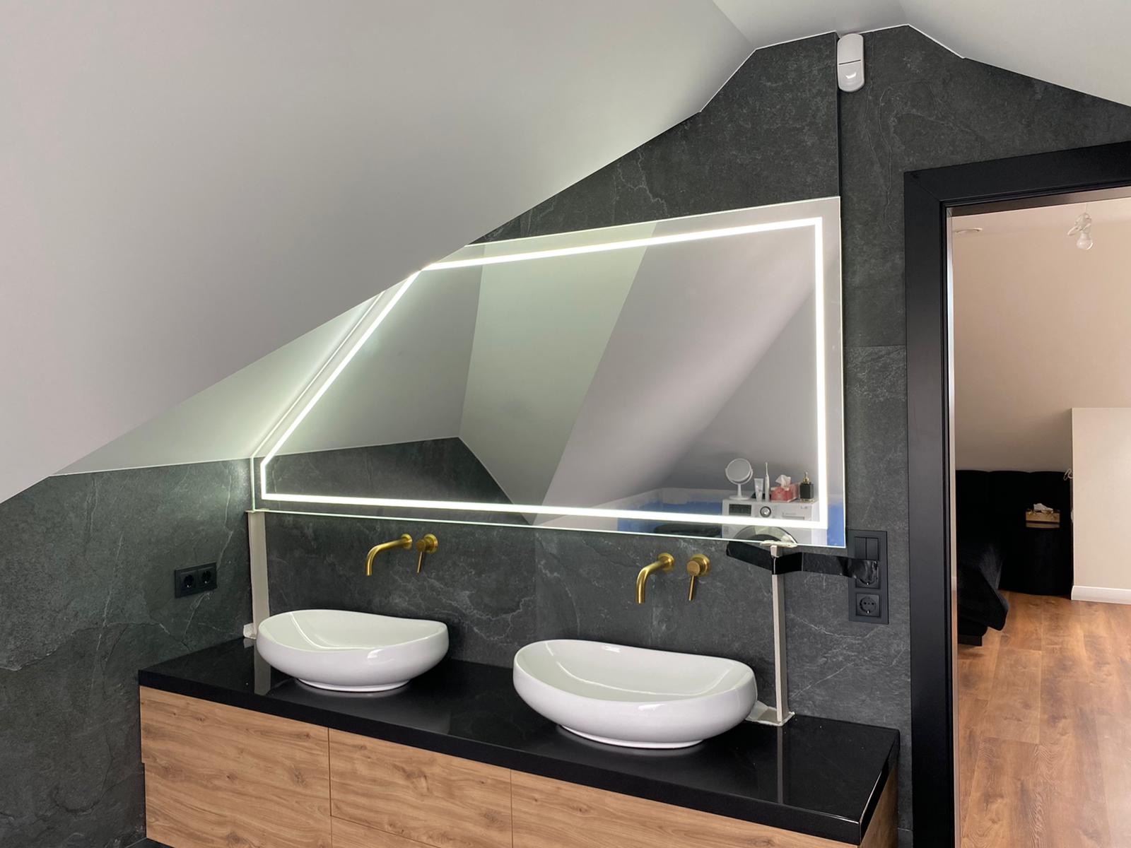 Nowoczesna łazienka na poddaszu z dwiema białymi umywalkami nablatowymi, złotą armaturą i podświetlanym lustrem w geometrycznej ramie, wykończona płytkami imitującymi kamień.
