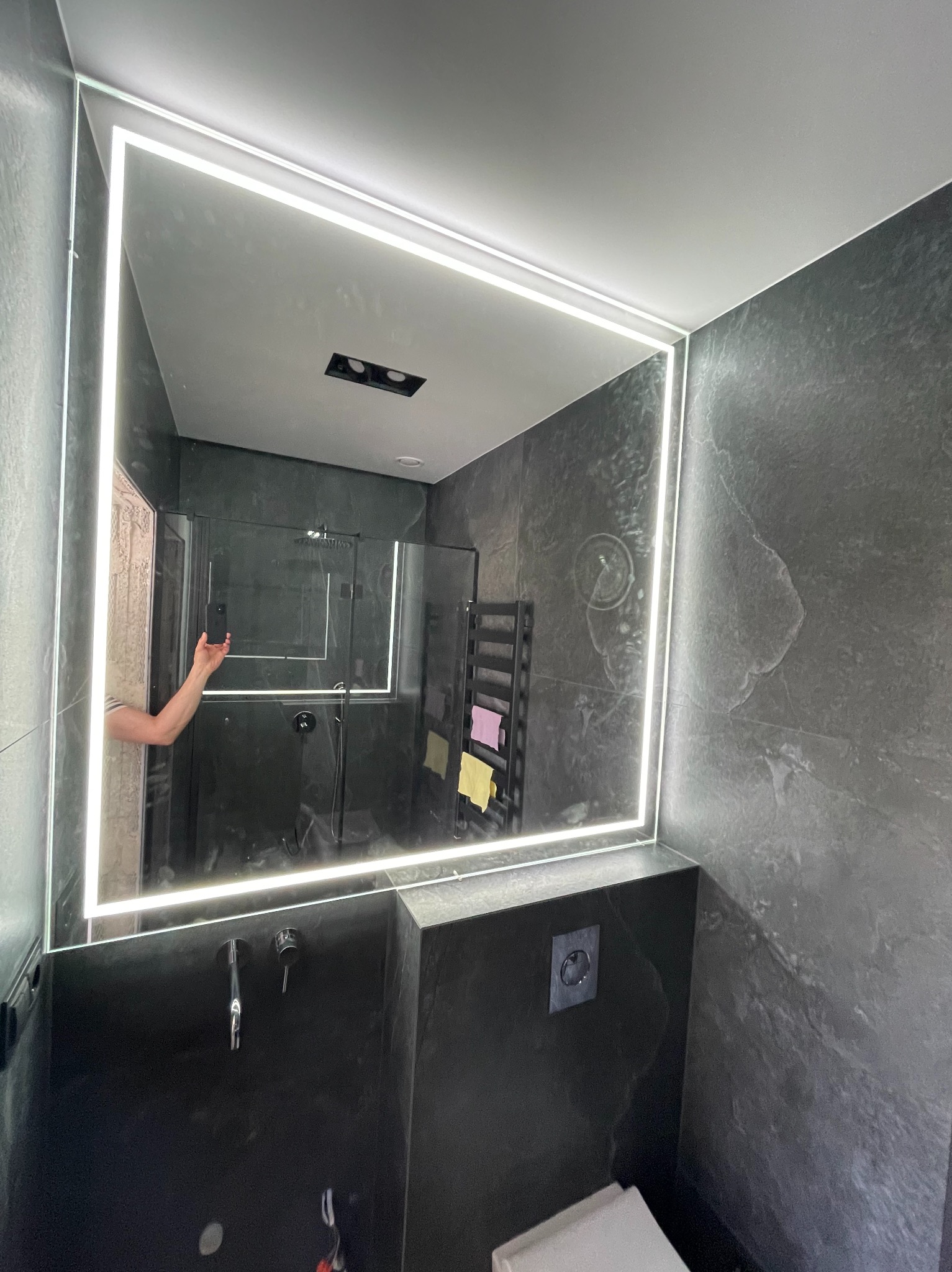 Nowoczesna łazienka z czarnymi płytkami, podświetlanym lustrem w kształcie kwadratu i szklaną kabiną prysznicową w tle. Widoczne elementy armatury i toaleta.