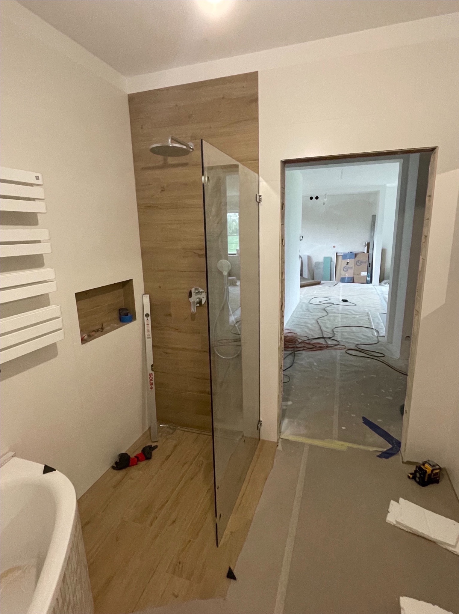 Wnętrze łazienki w trakcie remontu: zamontowana szklana kabina prysznicowa z deszczownicą, drewnopodobne płytki na ścianie i podłodze, grzejnik drabinkowy, wnęka w ścianie, poziomica oparta...