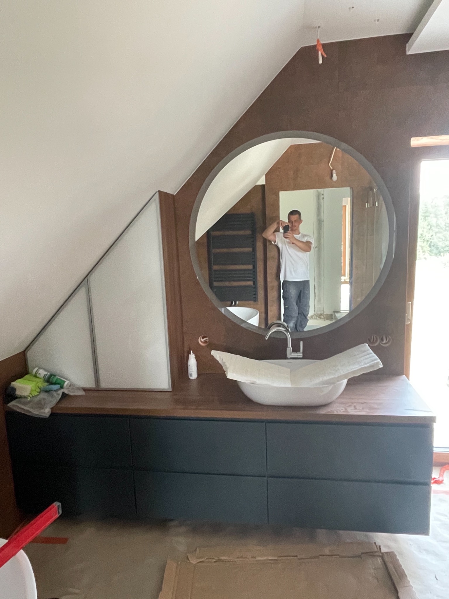 Łazienka na poddaszu w trakcie wykańczania: okrągłe lustro, umywalka nablatowa z baterią, brązowe płytki na ścianie, widoczne narzędzia i zabezpieczenia.