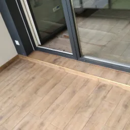 Wykończony balkon z drewnopodobnymi płytkami ceramicznymi ułożonymi w jodełkę, widok z góry na przesuwne okno balkonowe z czarną ramą.