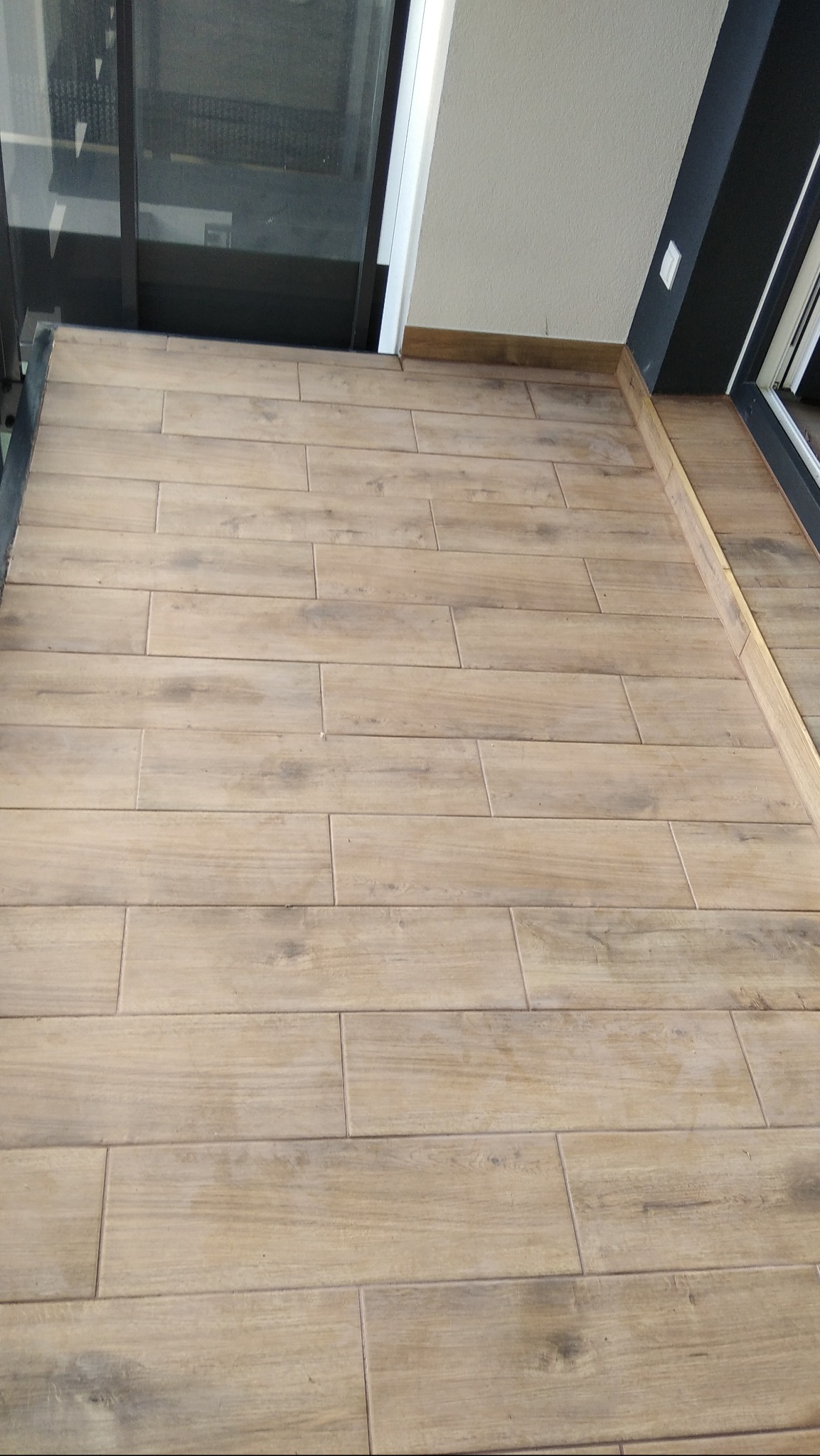 Balkon wyłożony podłużnymi, drewnopodobnymi płytkami gresowymi w odcieniach beżu i brązu, z widoczną ramą okienną i fragmentem ściany.