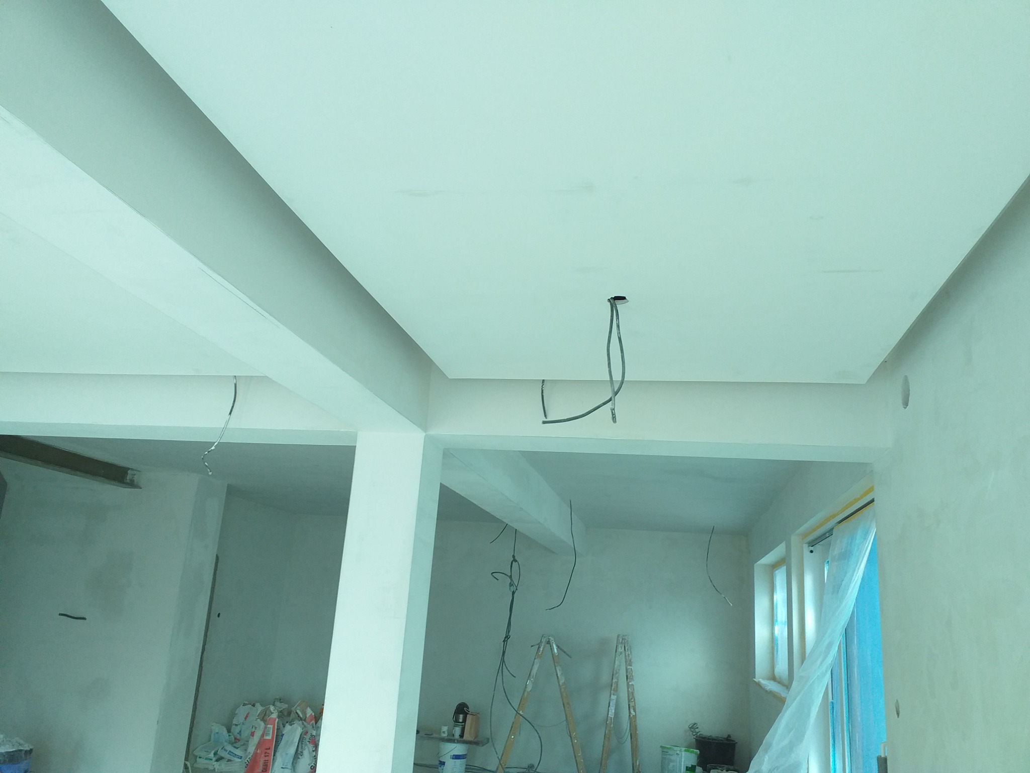 Wykończone sufity podwieszane z wystającymi przewodami elektrycznymi w surowym wnętrzu biurowym przed malowaniem, widoczne worki z zaprawą i drabina.